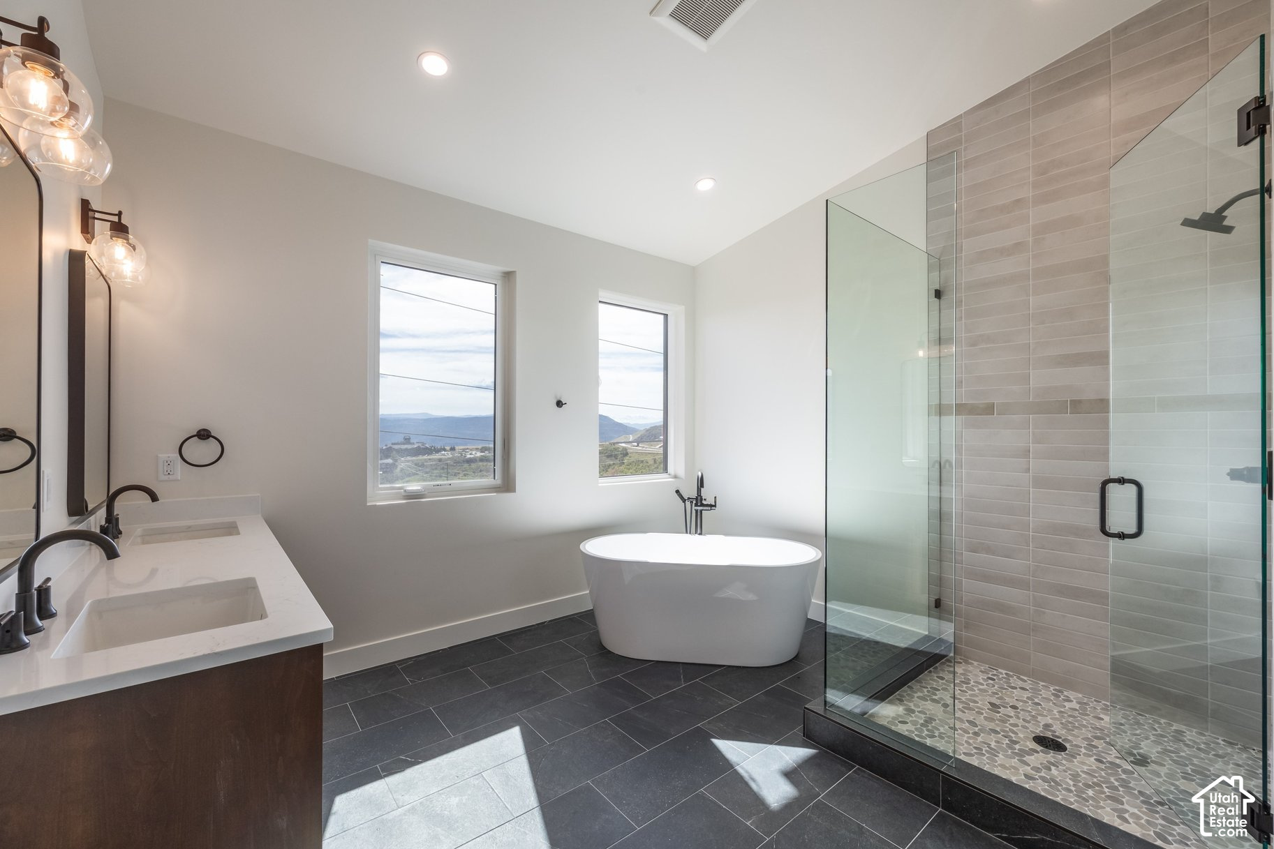 Modern bathroom with a freestanding tub, glass shower, double vanity, and mountain views through two windows.