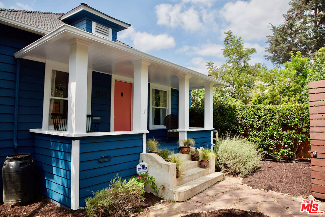 Blue house with a white-columned porch, coral-red door, and potted plants by the front steps.