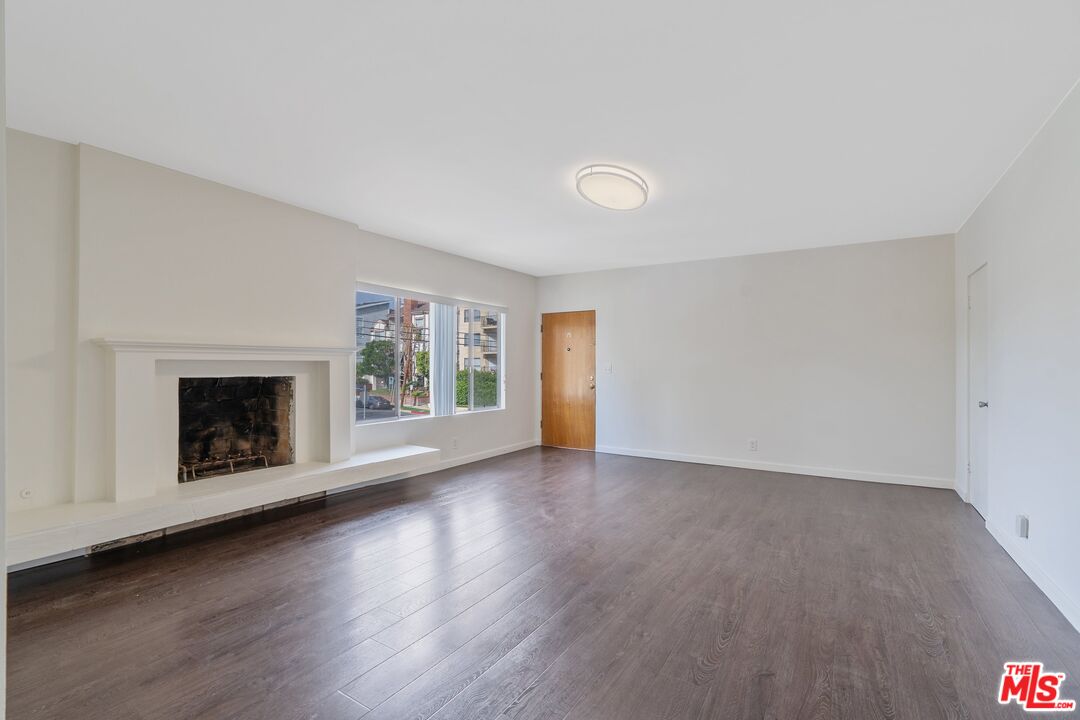 Vacant living room with dark wood floors, a white fireplace, and large windows.