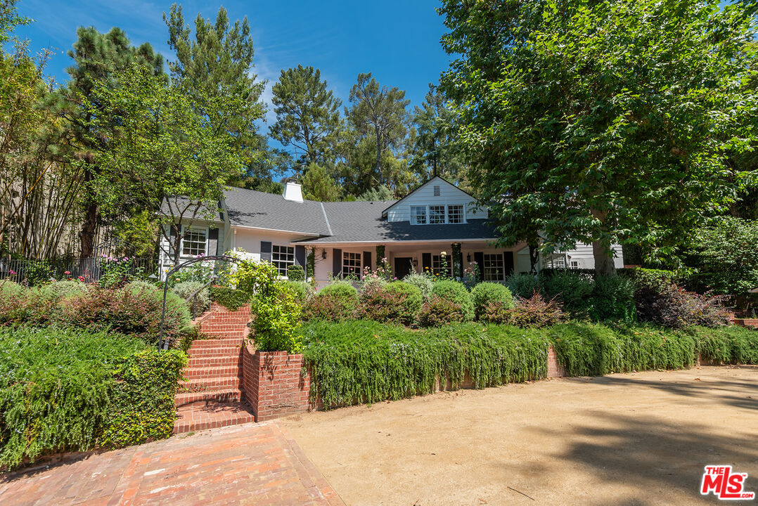 White house with a shaded porch, brick steps, and lush landscaping beneath mature trees.