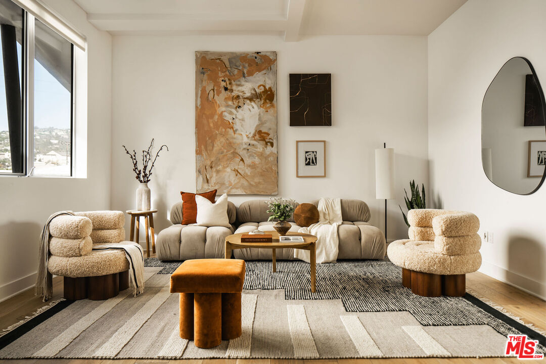 Staged living room with a rounded beige sofa, boucle armchairs, abstract art, and a large hillside-view window.