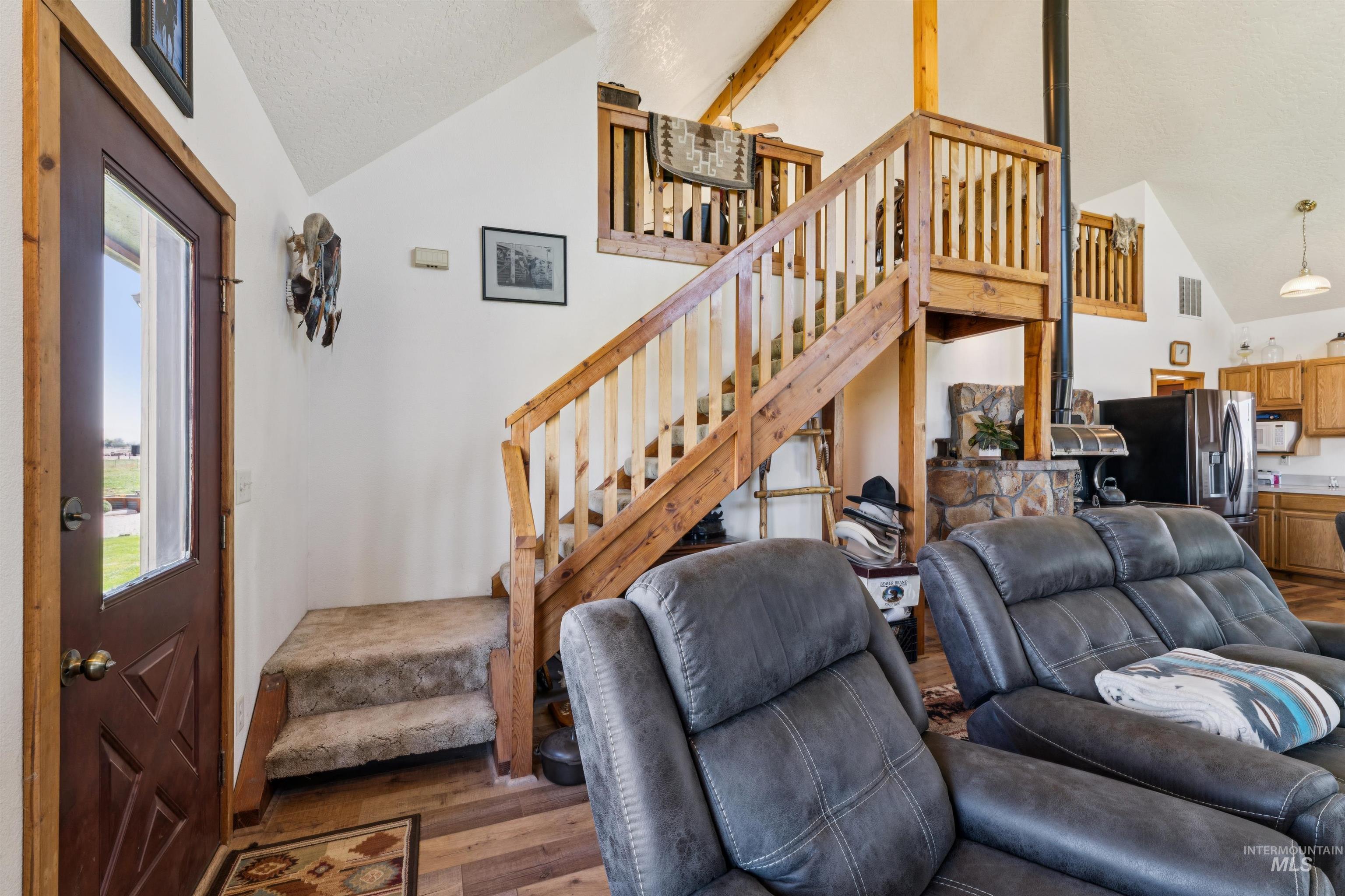 Living room with gray leather recliners, wooden staircase to a loft, and stone fireplace beside the kitchen.
