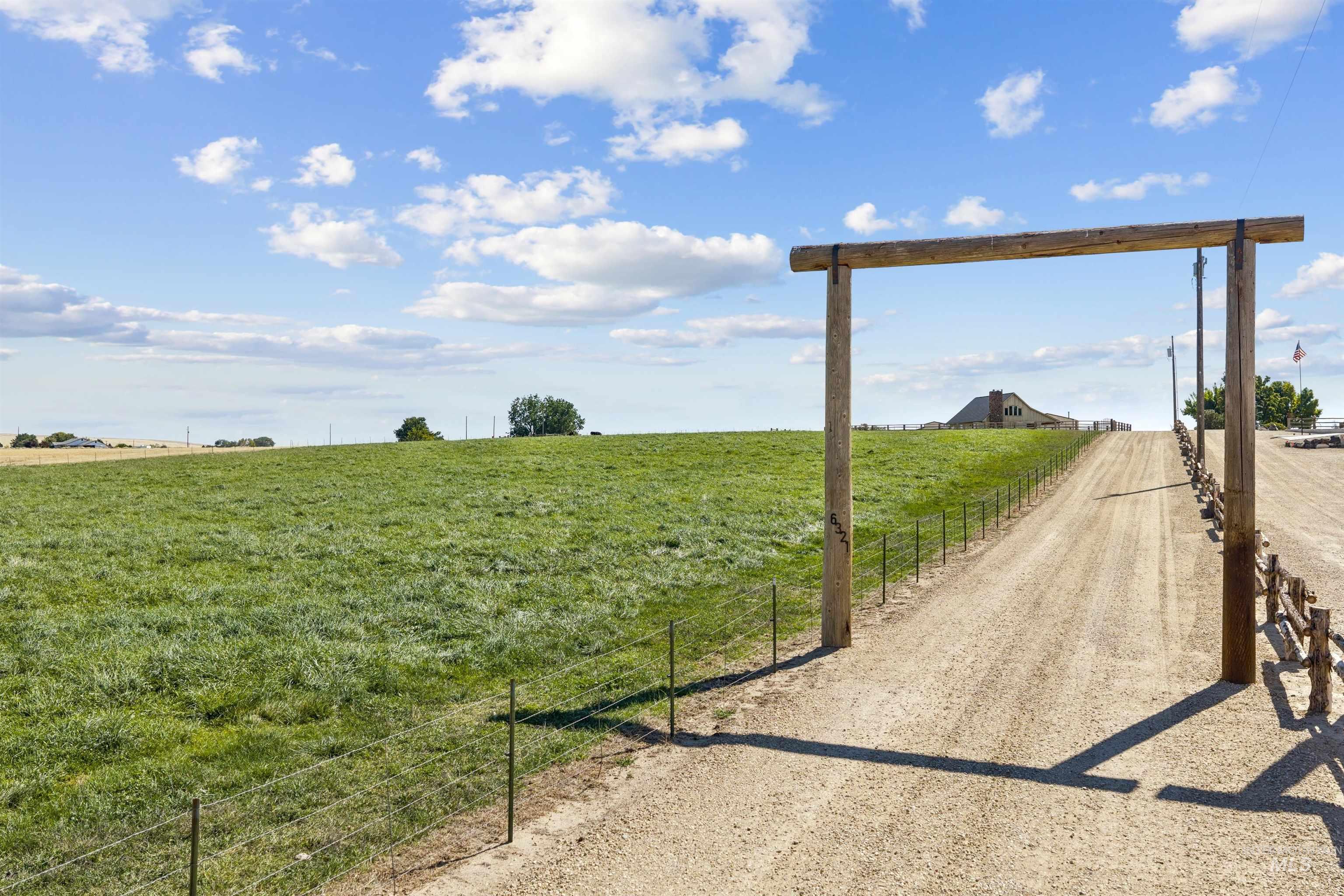 Rural property entrance with a wooden gateway, fenced grassy field, and gravel drive leading to a distant home.