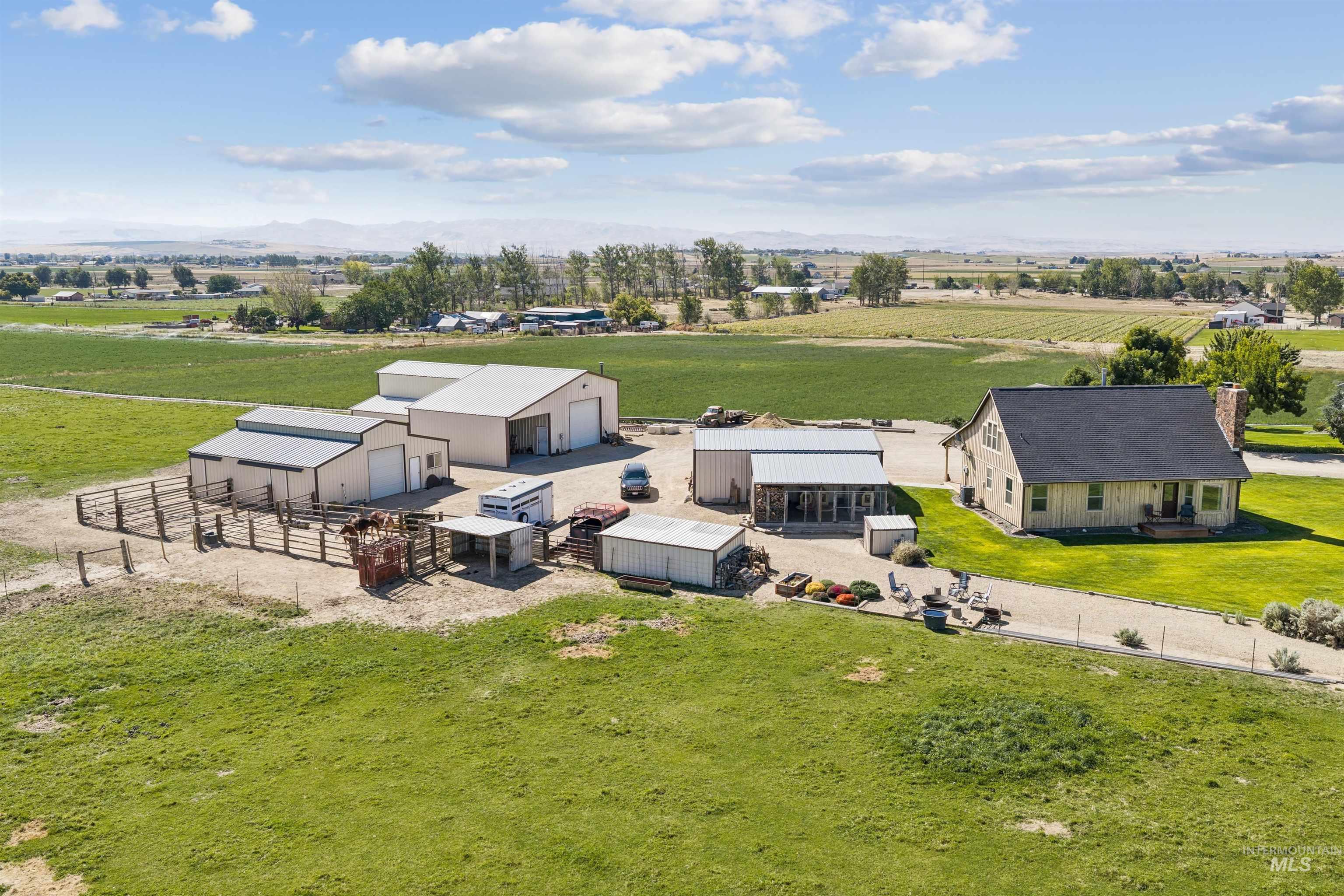 Aerial view of a farmhouse, metal barns, livestock pens, and green fields with distant mountains.