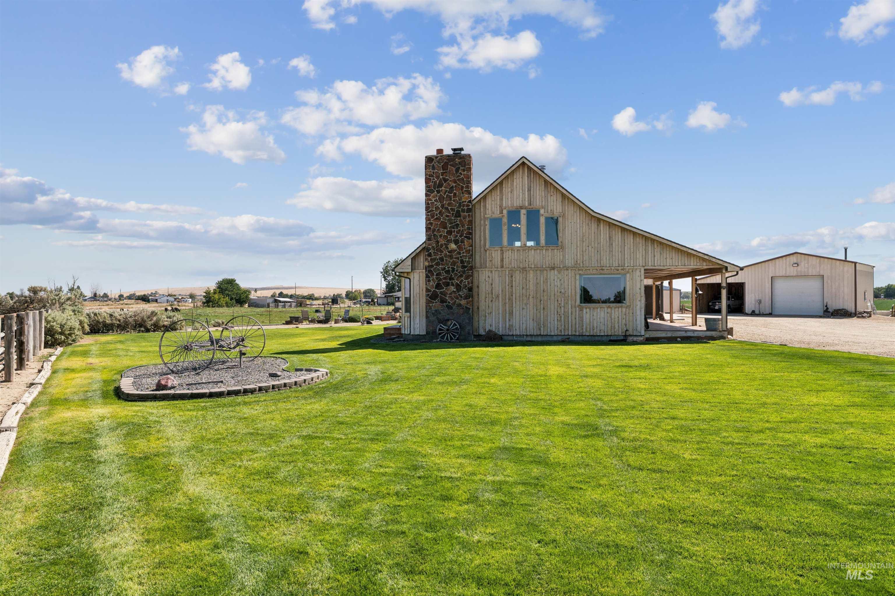 Wood-sided rural home with a stone chimney and detached garage behind a broad green lawn.