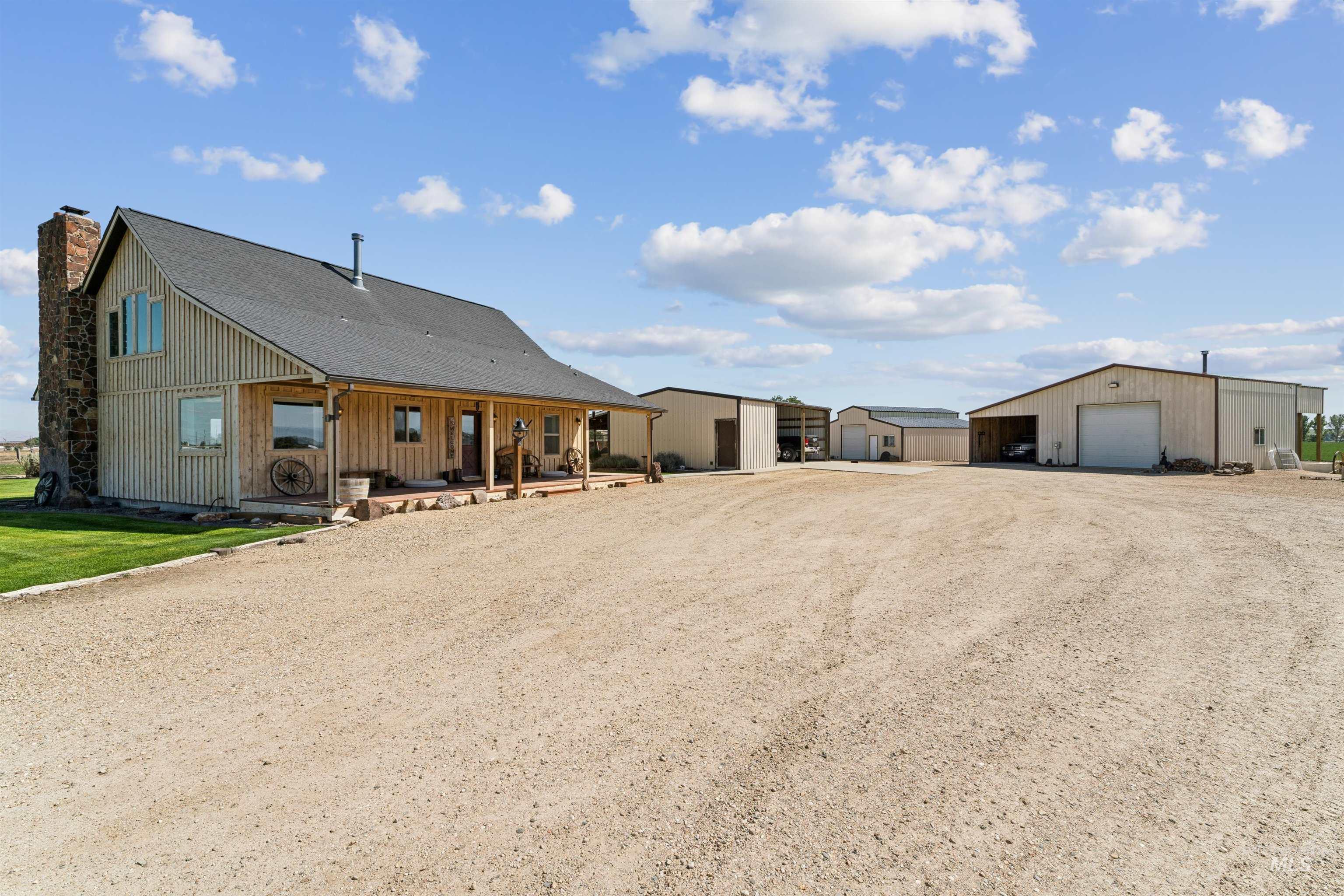 Wood-sided rural home with a stone chimney, covered porch, gravel driveway, and several detached outbuildings.