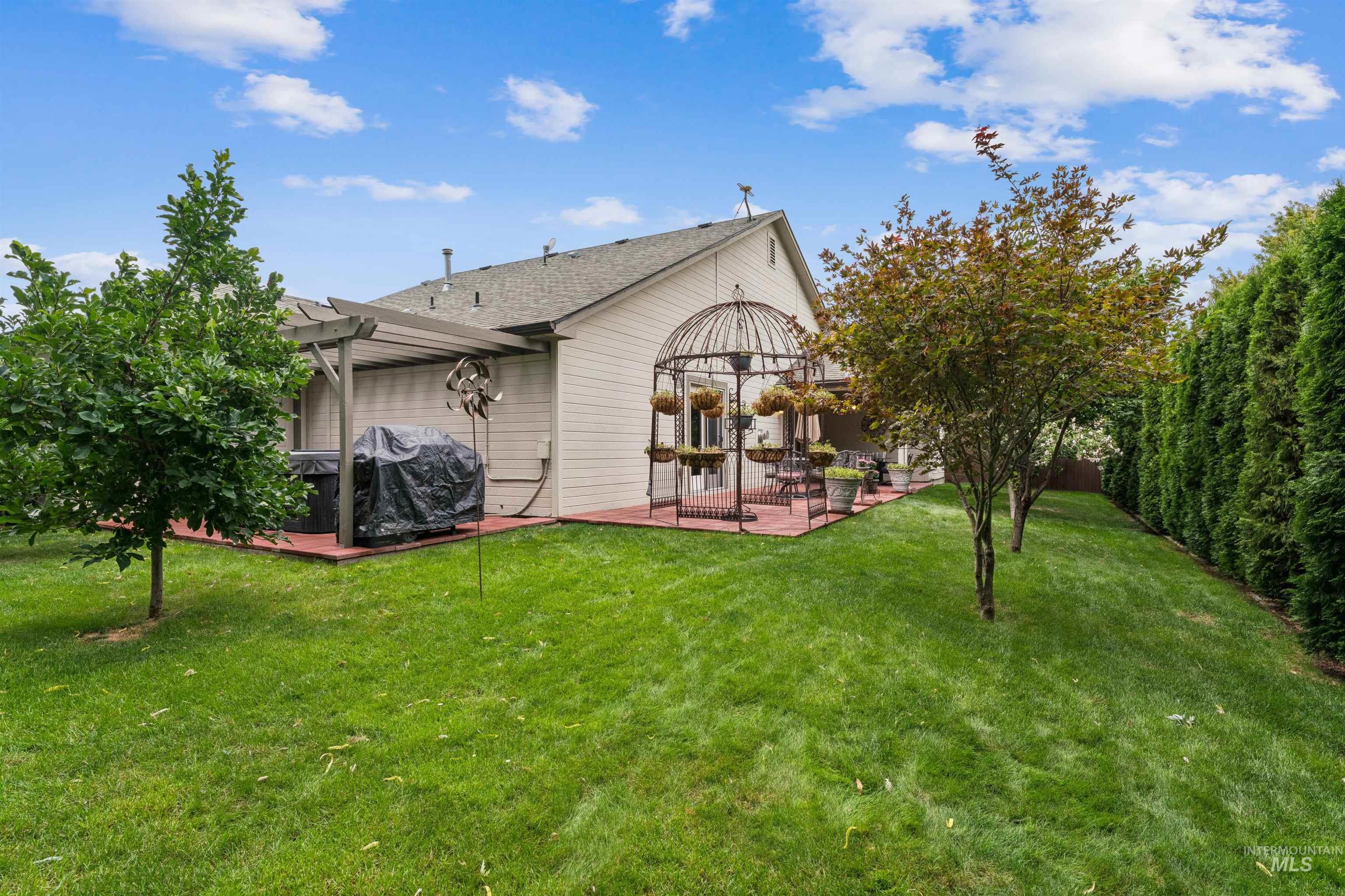 Home’s backyard with a patio arbor, covered pergola, broad lawn, ornamental trees, and tall privacy hedge.