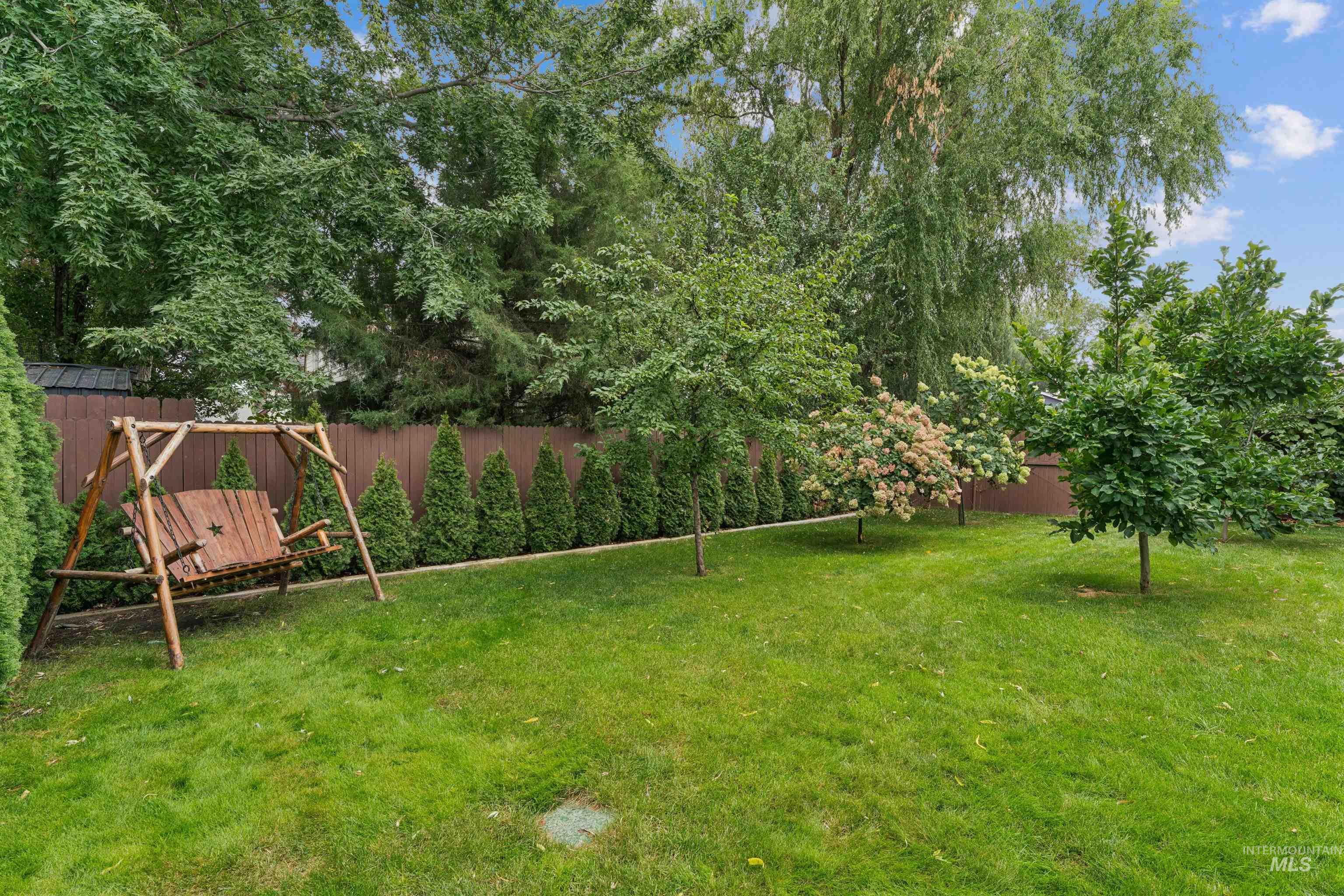 Backyard lawn with a wooden swing, mature trees, flowering shrubs, and a privacy fence.