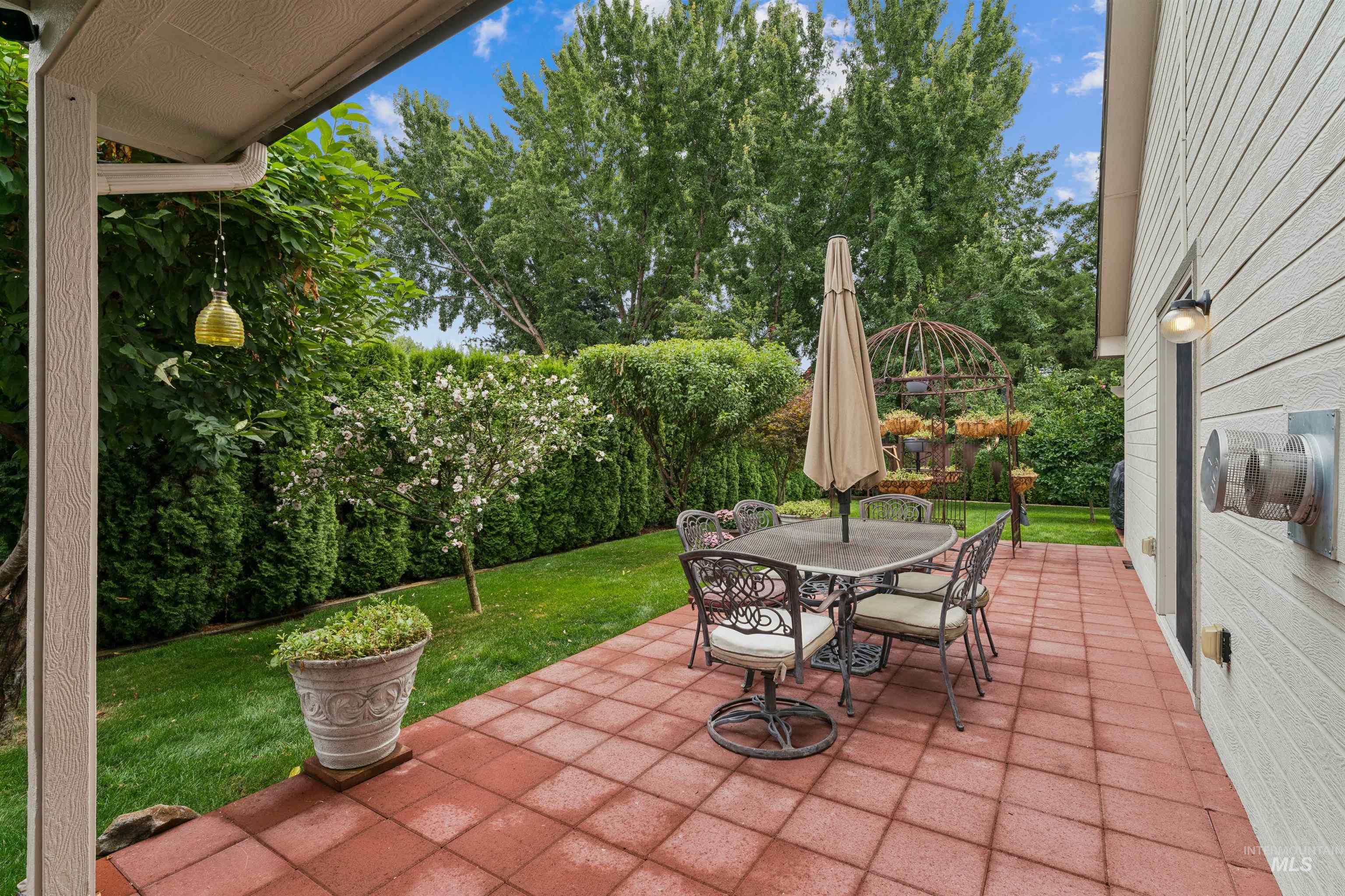 Backyard patio with red pavers, an outdoor dining set, flowering tree, and lush hedges.