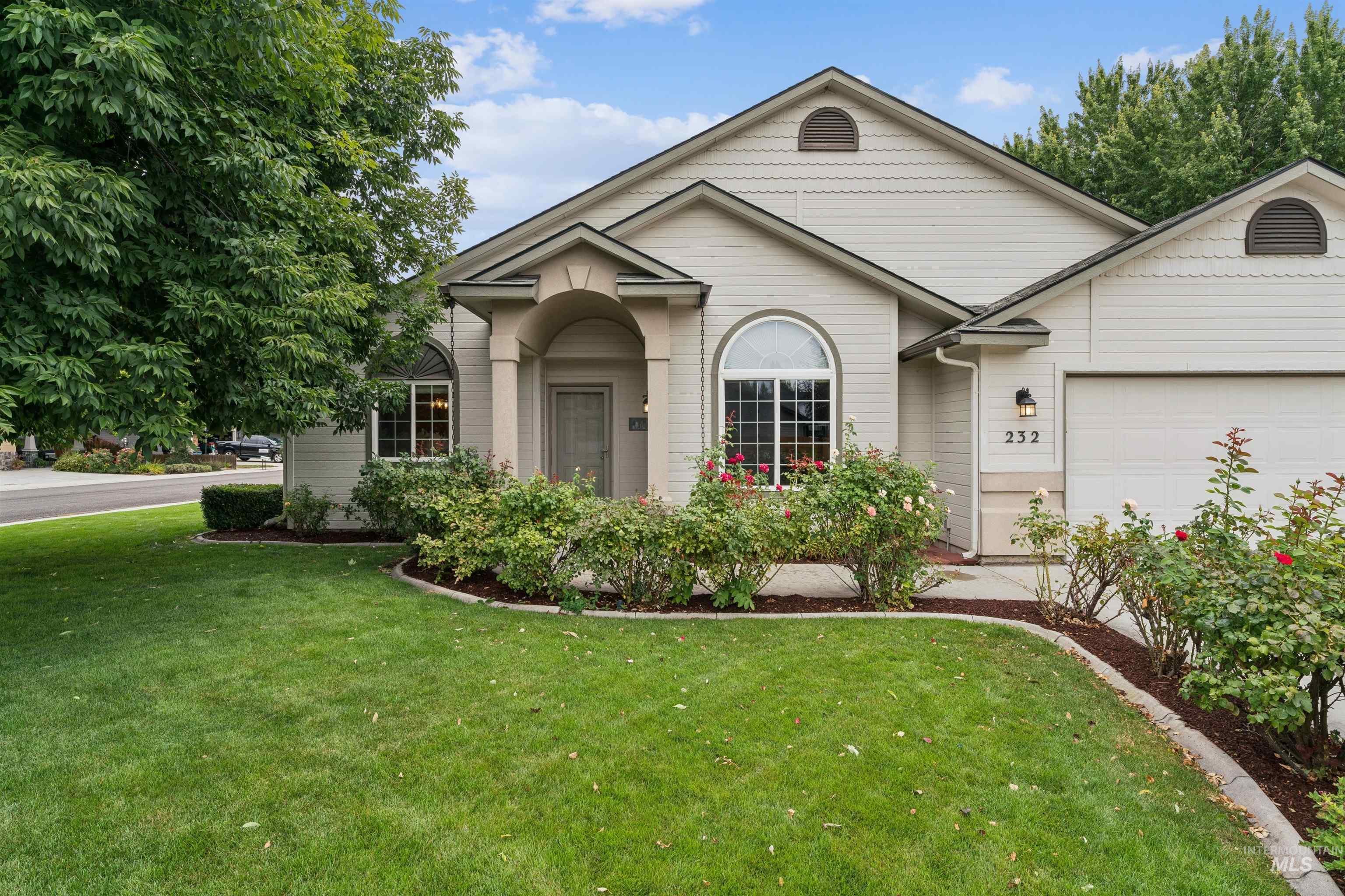 Beige house with an arched entry, attached garage, flowering shrubs, and green lawn.