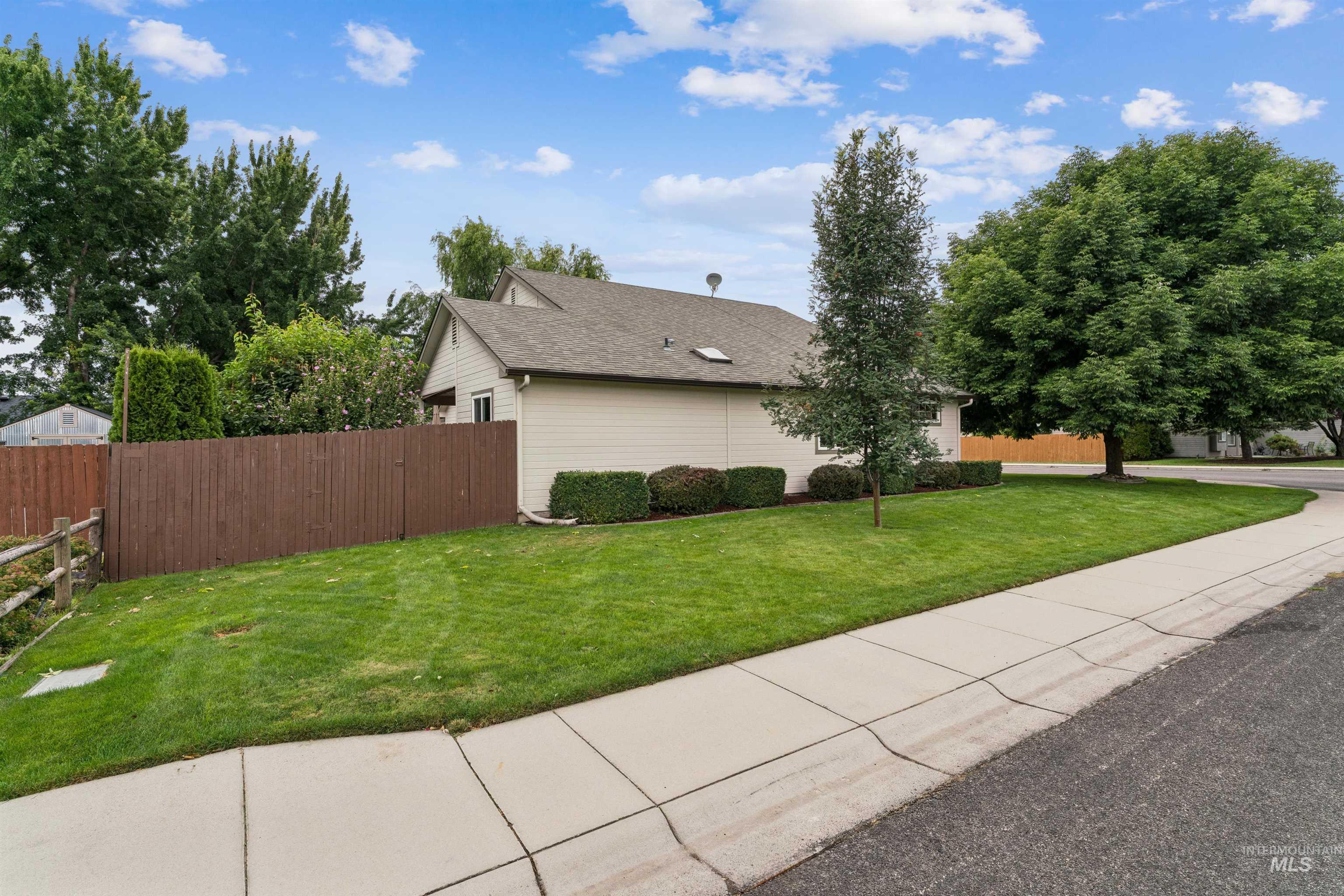 Light-colored house with a brown privacy fence, landscaped lawn, and mature trees beside the sidewalk.