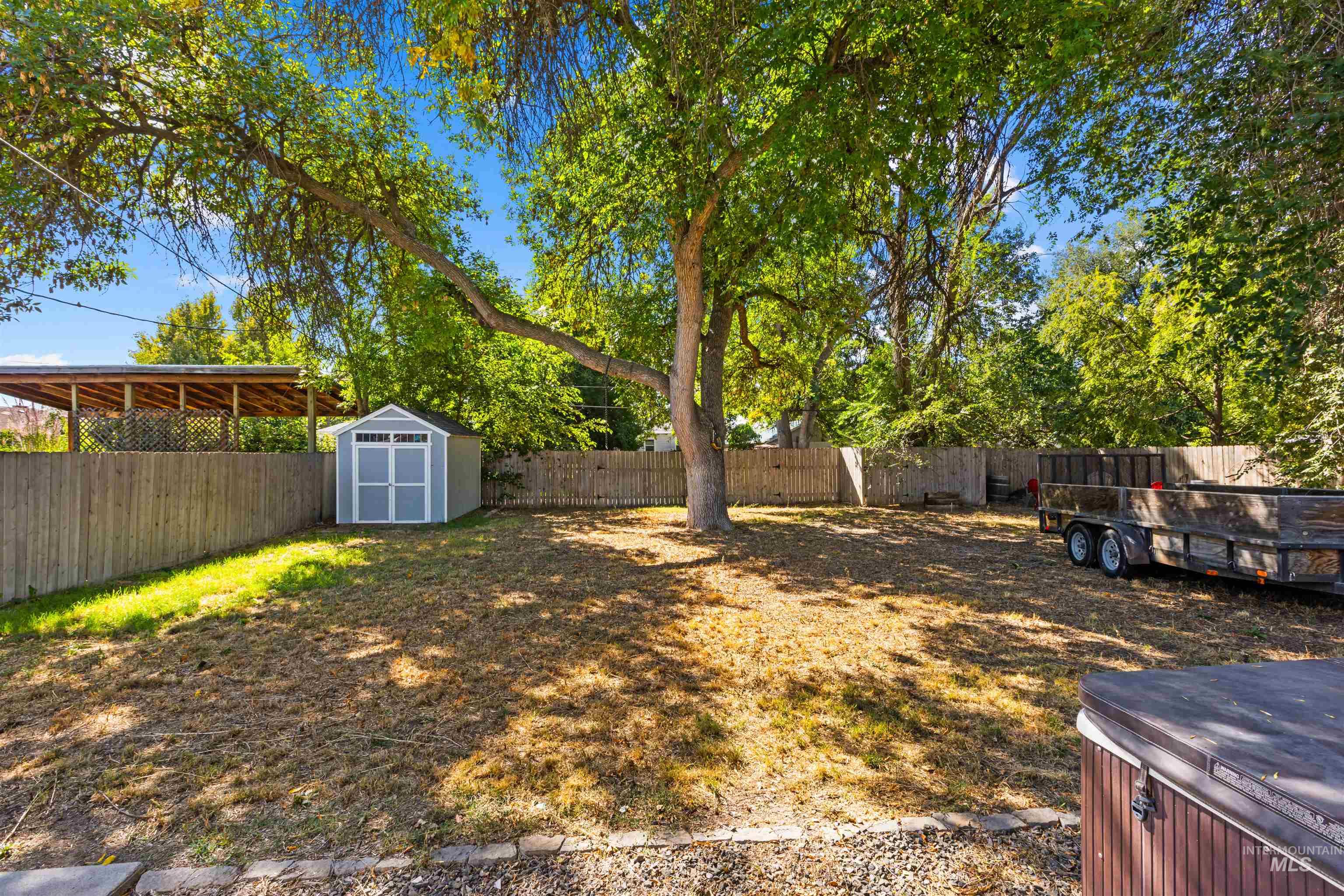 Fenced backyard shaded by mature trees, with a storage shed, covered patio, trailer, and hot tub.