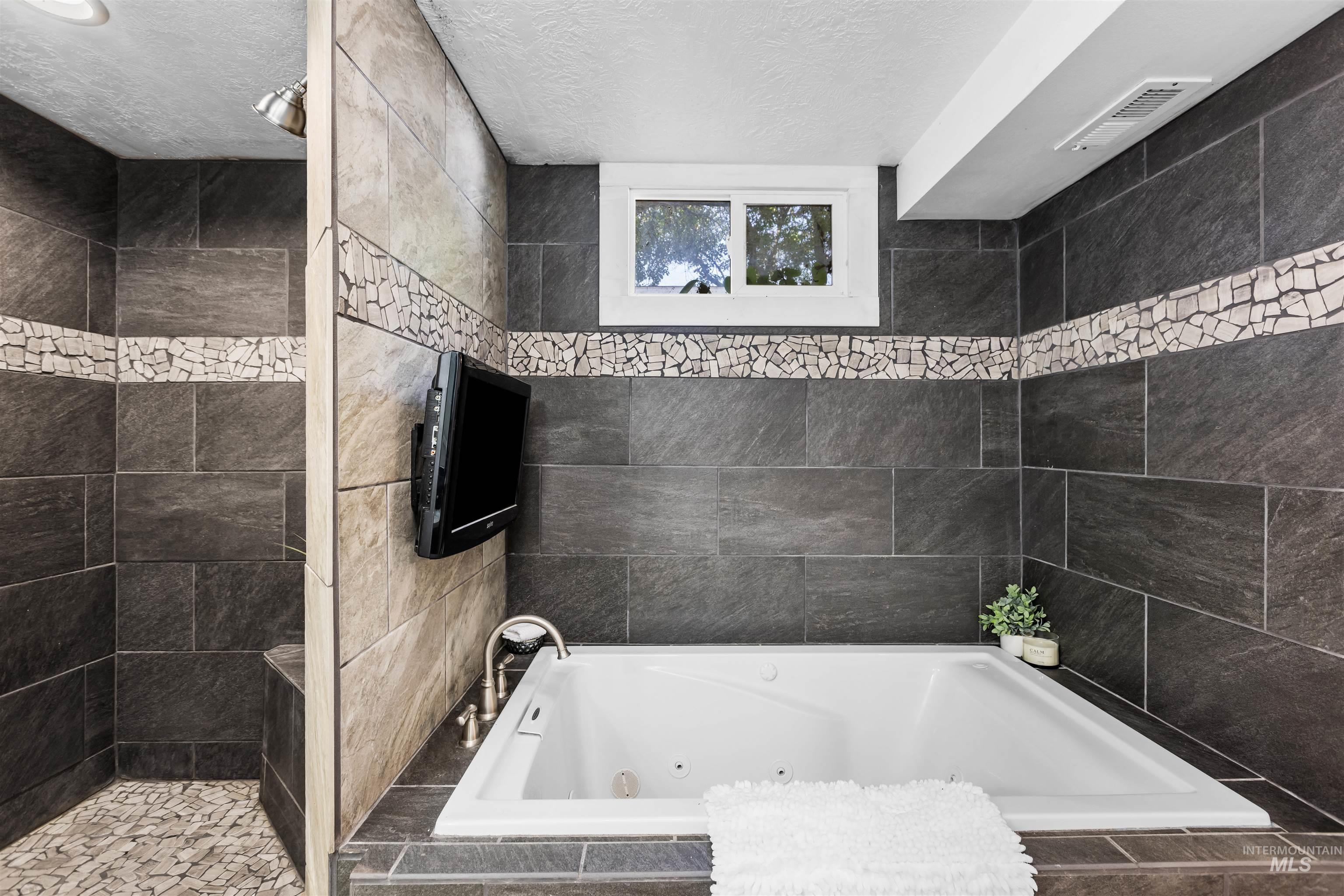 Bathroom with a large jetted tub, dark tile, mosaic accents, and a wall-mounted television.