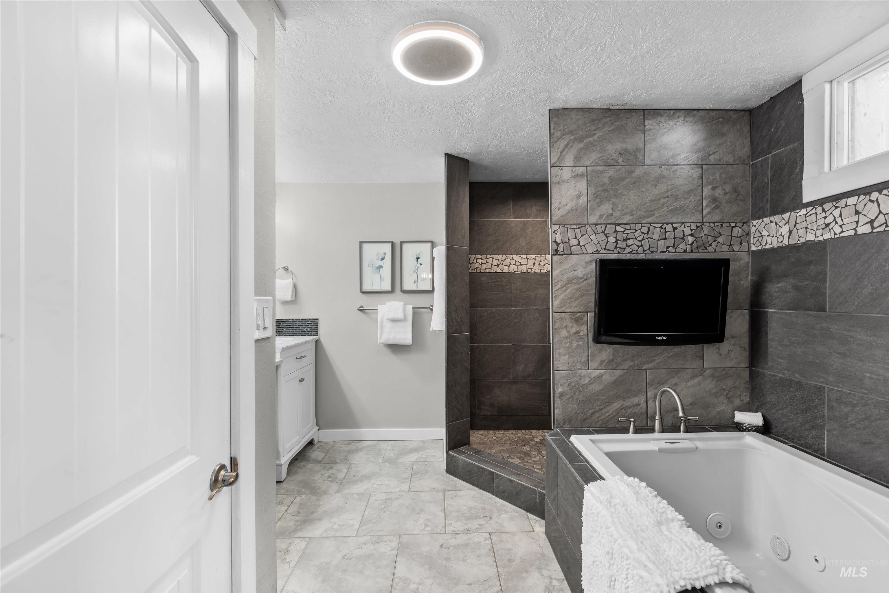 Bathroom with a built-in jetted tub, dark tiled shower, and wall-mounted television.