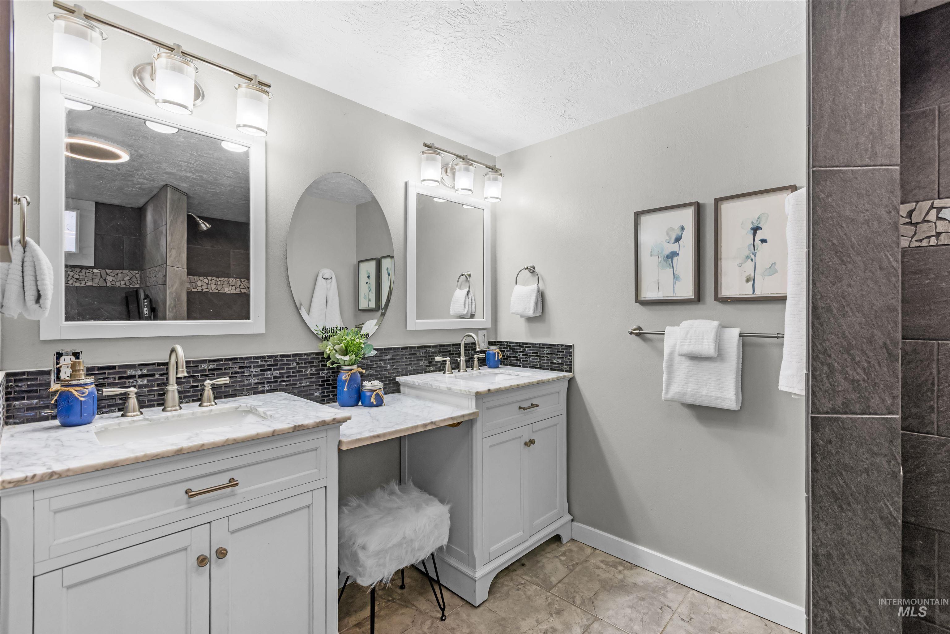 Bathroom with double white vanities, marble-look countertops, dark tile backsplash, and a tiled shower.