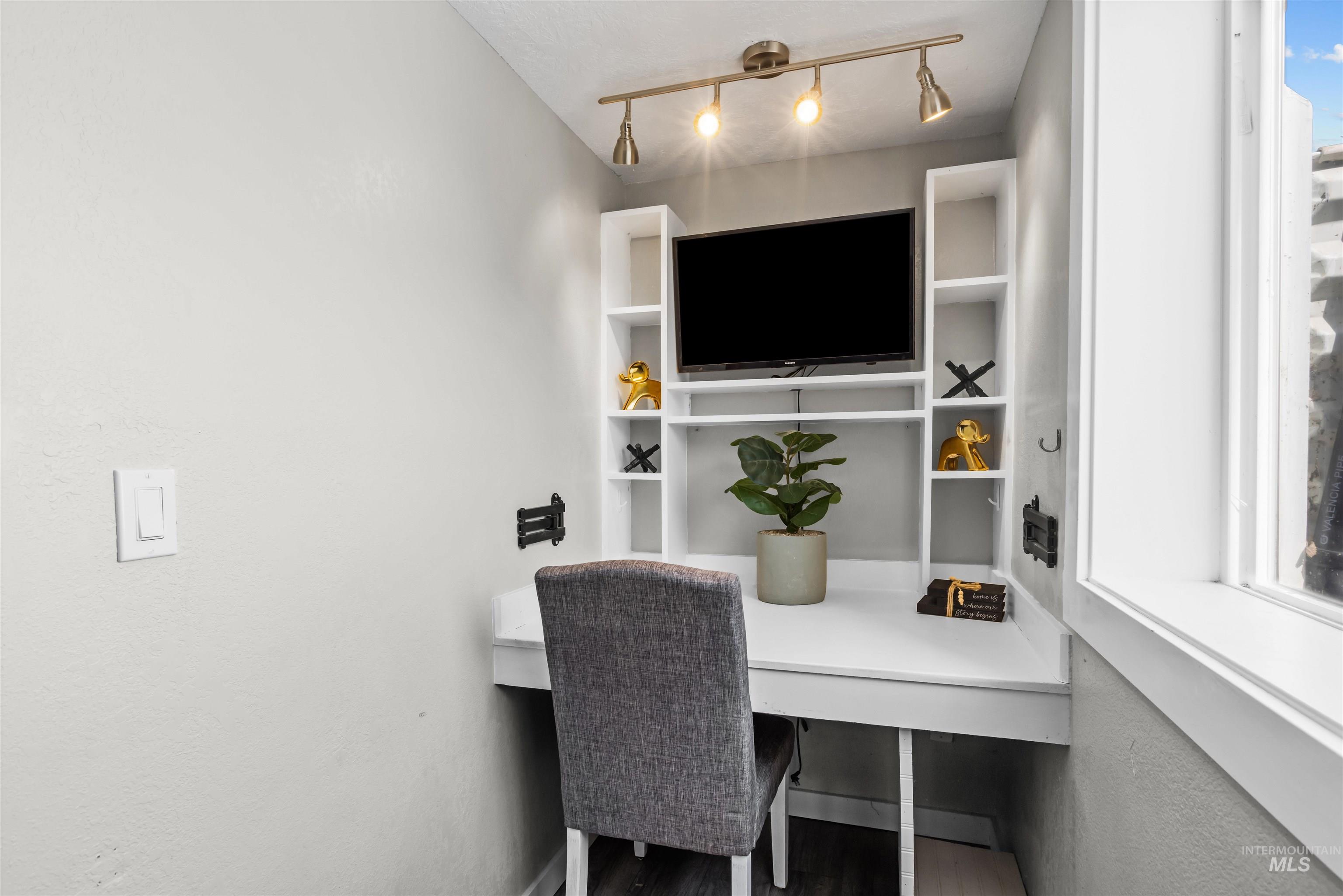 Home office nook with built-in desk and shelving, television, gray chair, and large window.