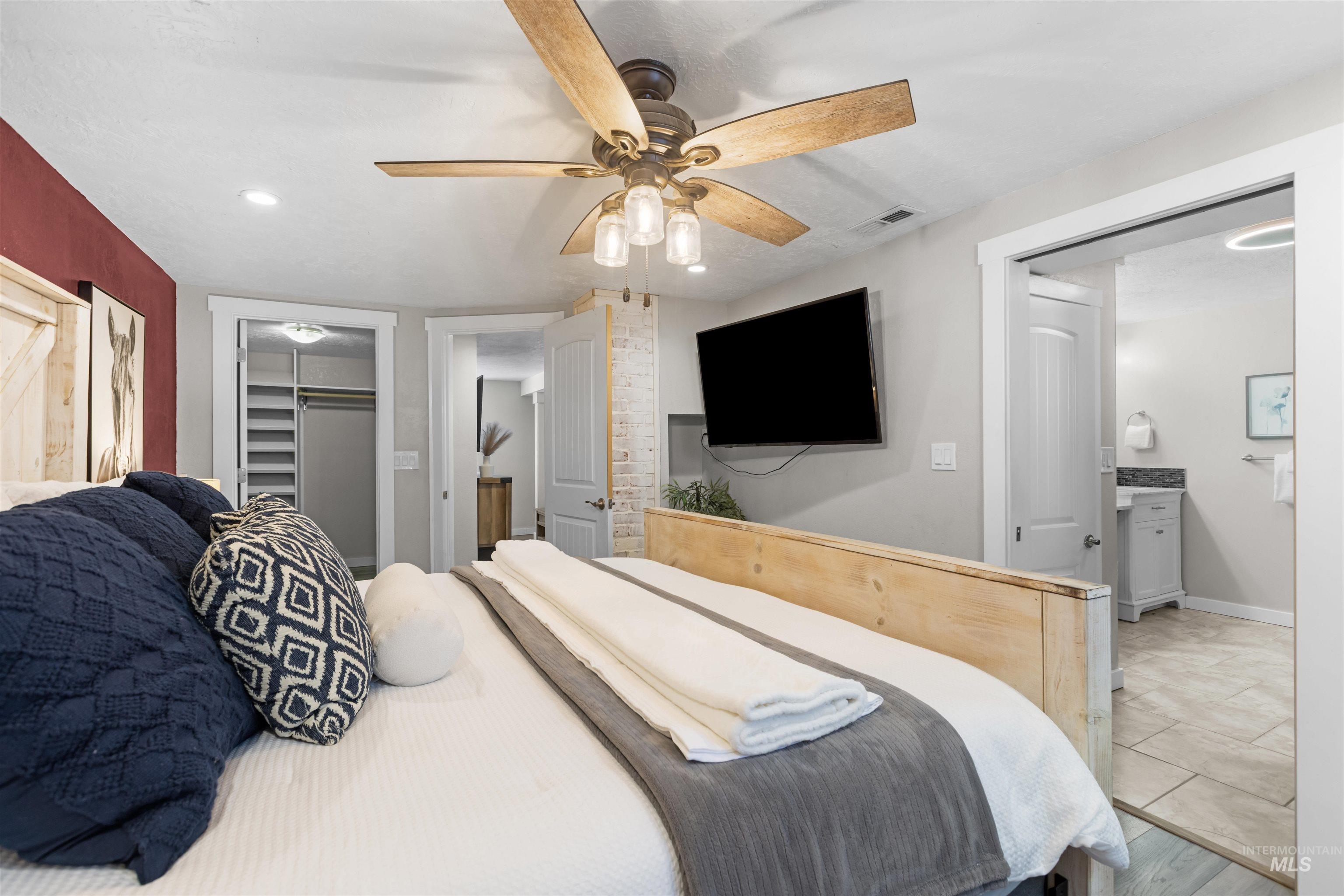 Staged bedroom with a white bed, wood ceiling fan, wall-mounted TV, and doorway to an adjoining bathroom.