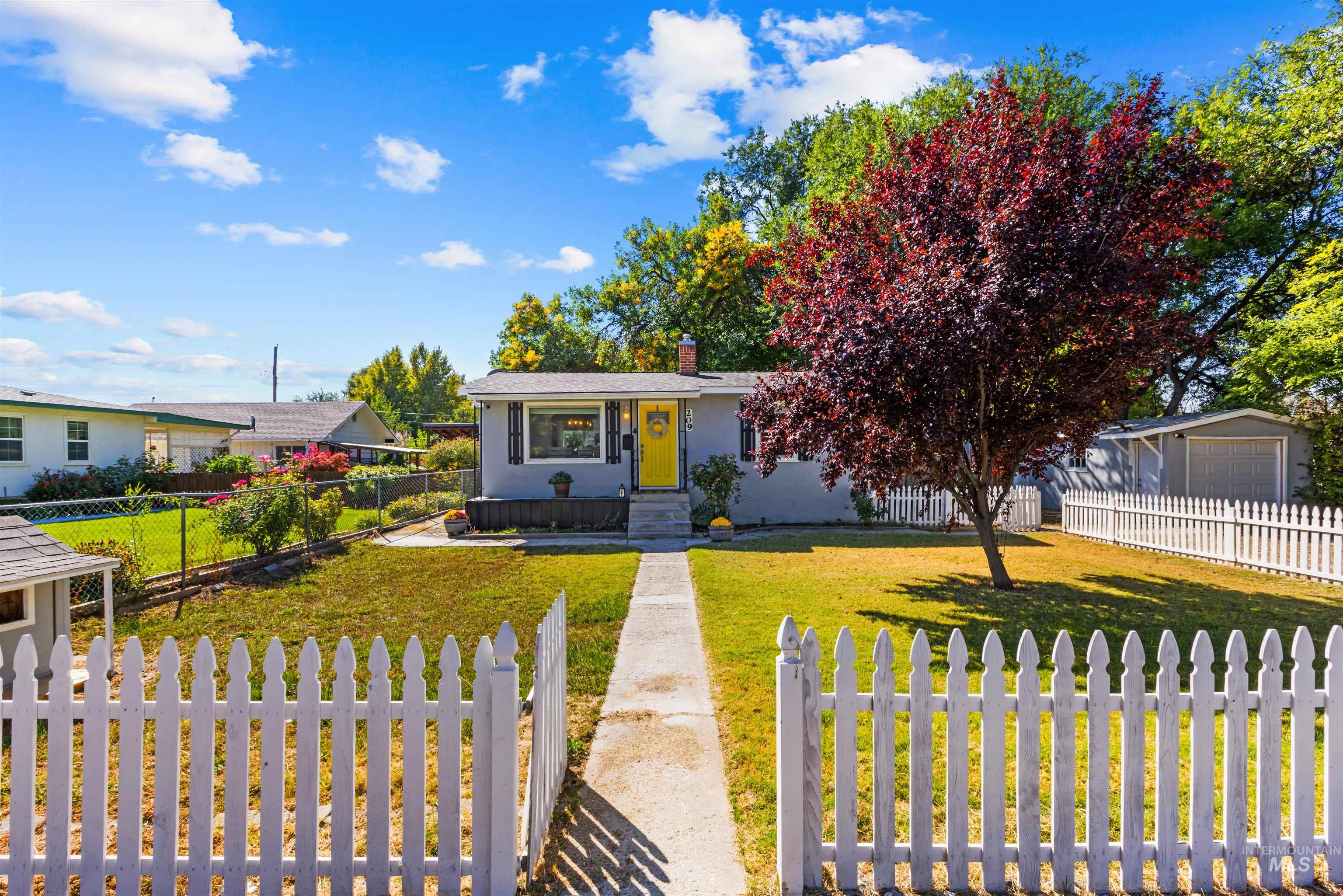 Single-story home with a yellow front door, white picket fence, and large red-leaf tree.