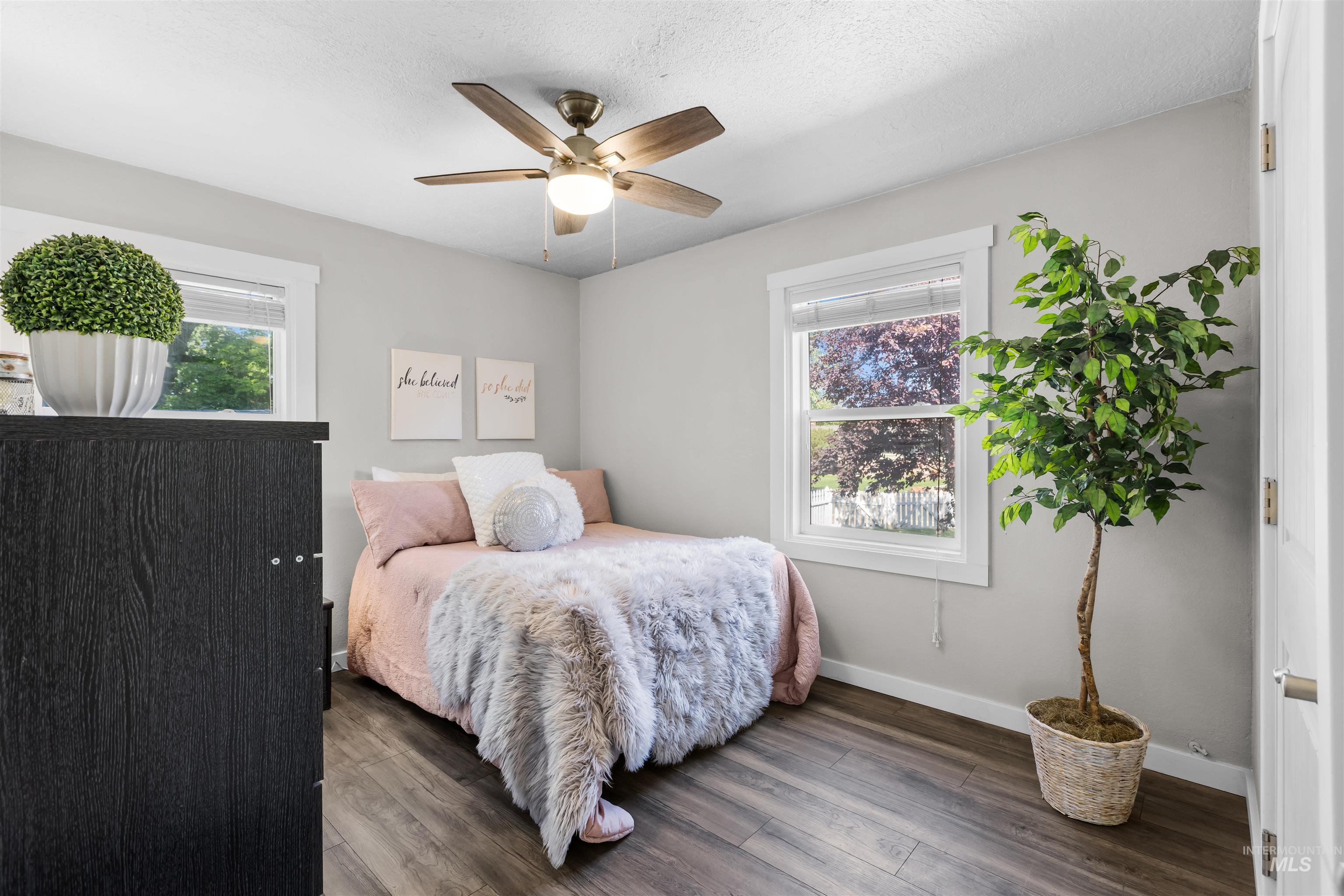 Staged bedroom with a blush-pink bed, fluffy throw, ceiling fan, and two windows.