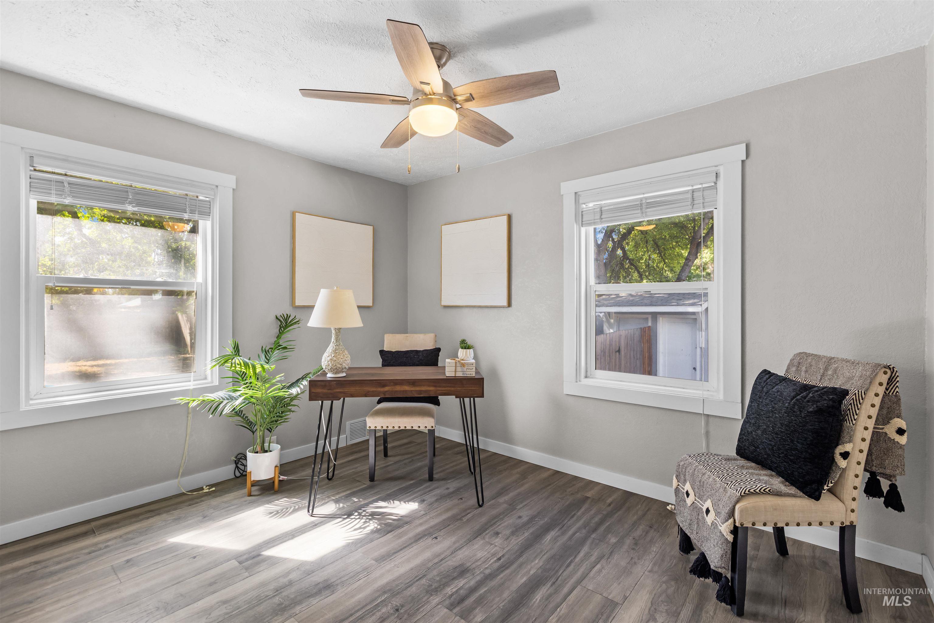 Staged home office with a wood desk, two windows, potted palm, and patterned armchair.