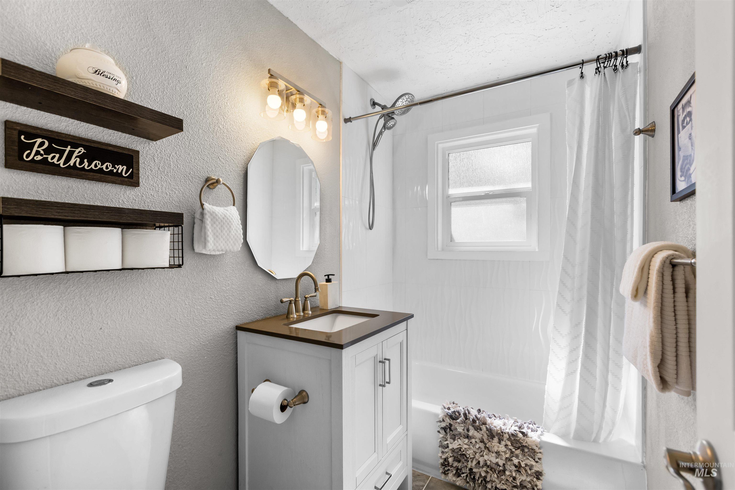 Bathroom with a white tub and shower, window, and vanity with dark countertop and brass fixtures.