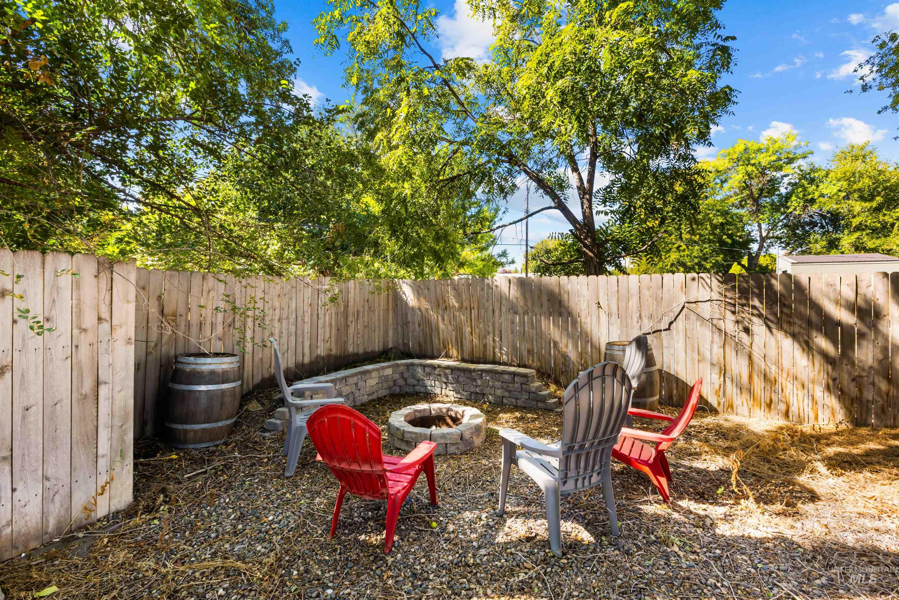 Fenced backyard with colorful chairs around a stone fire pit beneath mature trees.