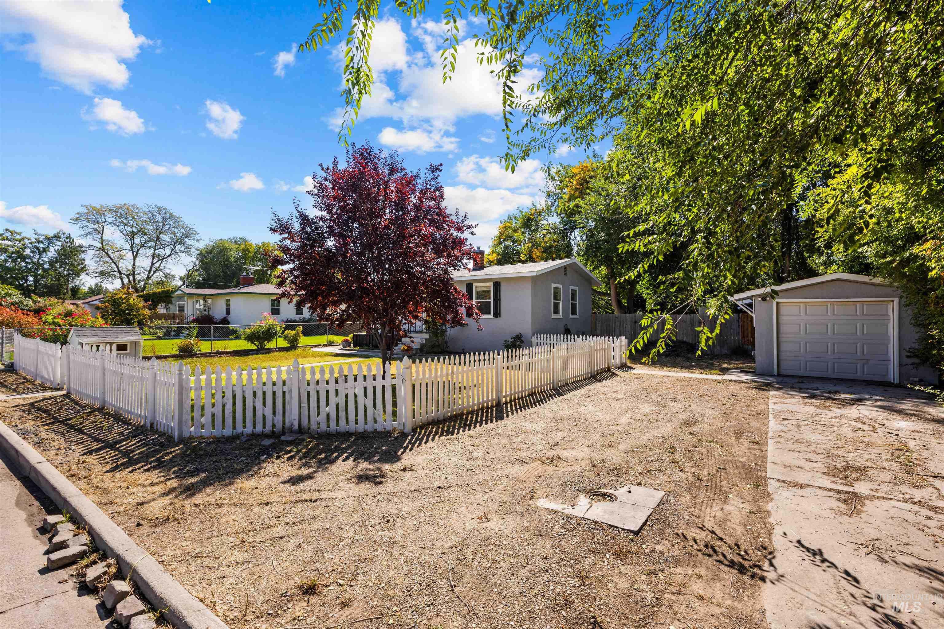 Gray single-story home behind a white picket fence, with mature trees and a detached garage.