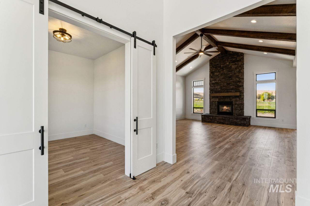 Living room with a stone fireplace and exposed wood beams, viewed beside an empty room through a barn door.