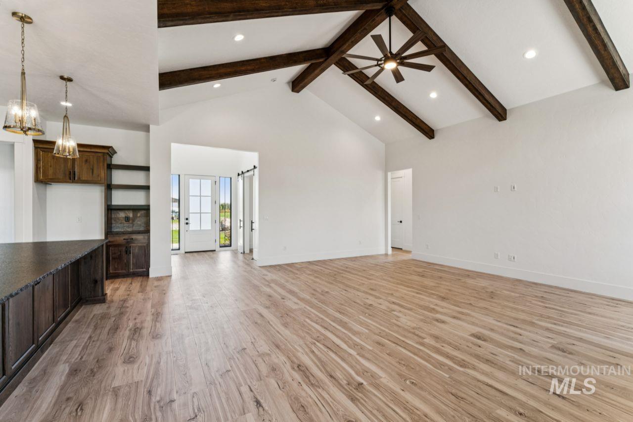 Empty open-plan living room with vaulted ceiling, dark exposed beams, and light wood-look floors.