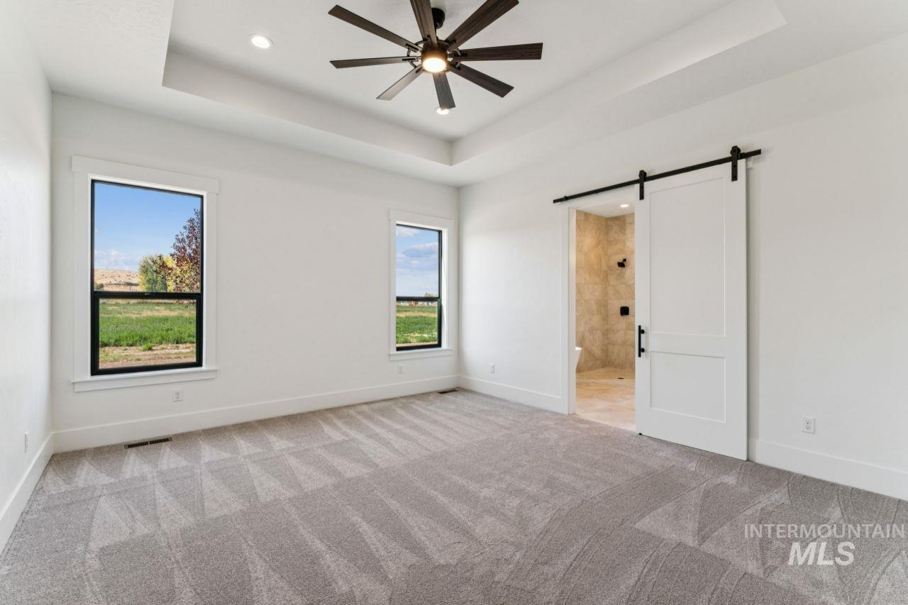 Empty carpeted bedroom with two windows, a ceiling fan, and an open barn door to a tiled bathroom.