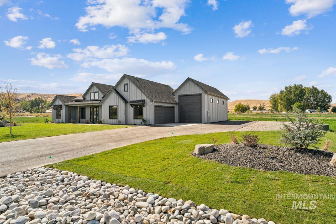 Gray gabled home with a tall garage, broad driveway, landscaped lawn, and low hills in the distance.