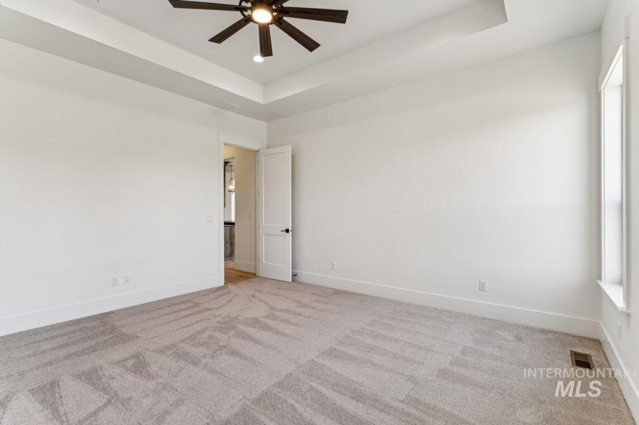 Empty carpeted bedroom with a tray ceiling, dark ceiling fan, and tall window.