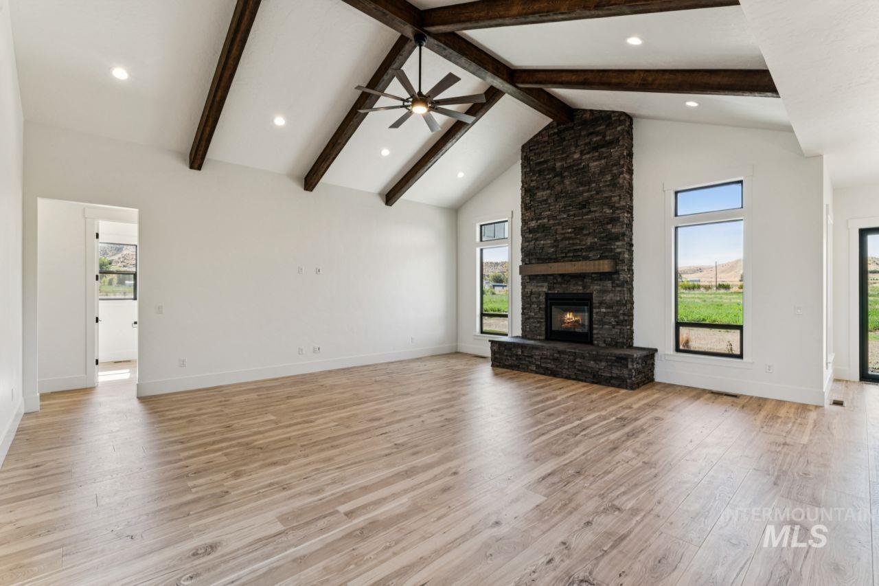 Empty living room with a tall stone fireplace, exposed wood beams, and broad windows overlooking greenery.