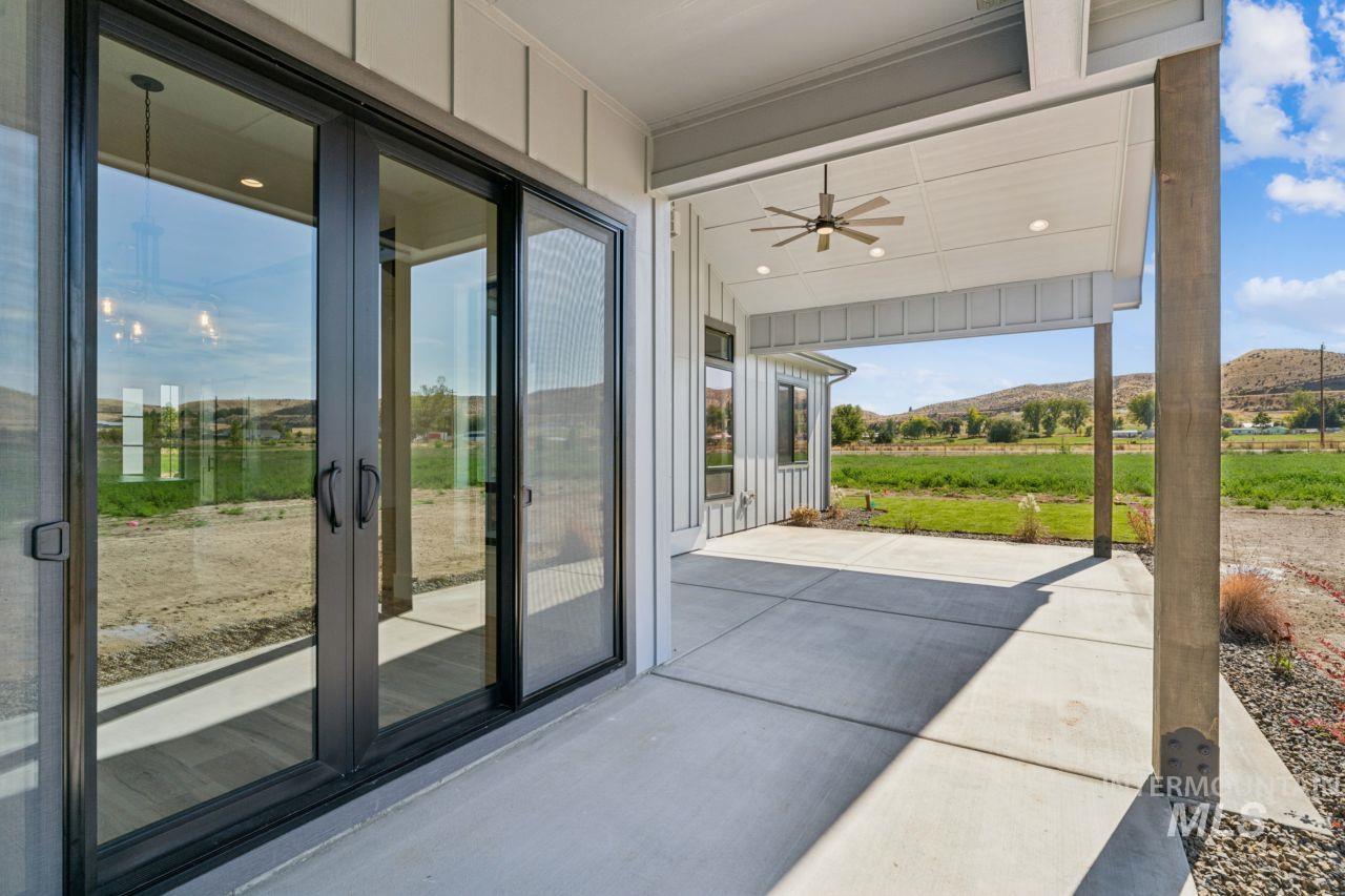Covered concrete patio with sliding glass doors and a ceiling fan, overlooking open fields and low hills.