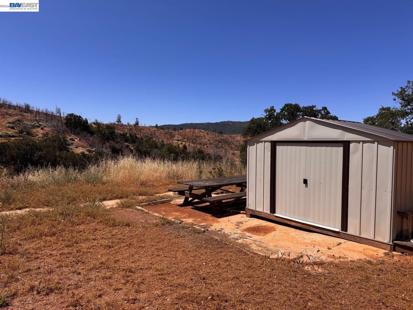 Metal storage shed and picnic table on dry acreage with rolling hills in the distance.