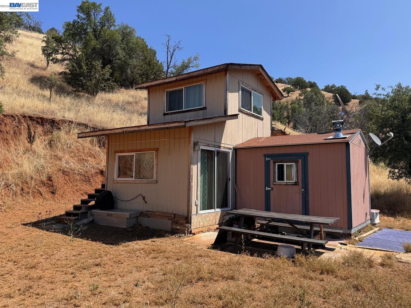 Tan rural home with attached red outbuilding and picnic table on a dry hillside.