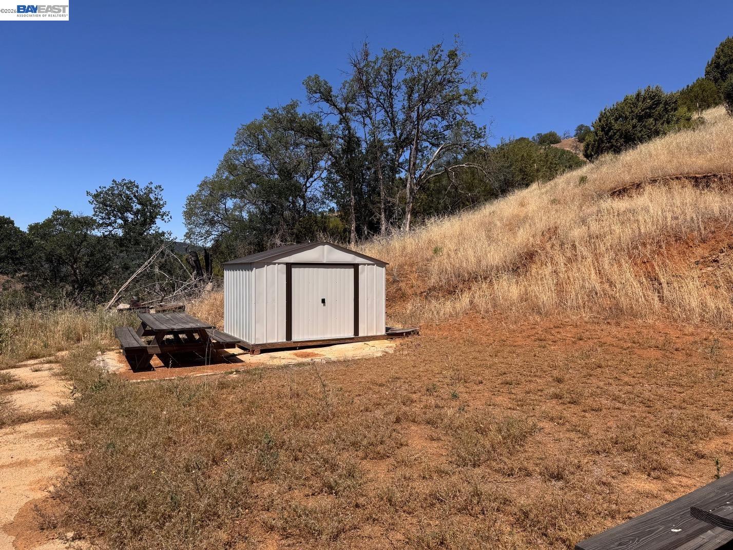 White metal shed and picnic tables on a dry hillside among trees and golden grass.