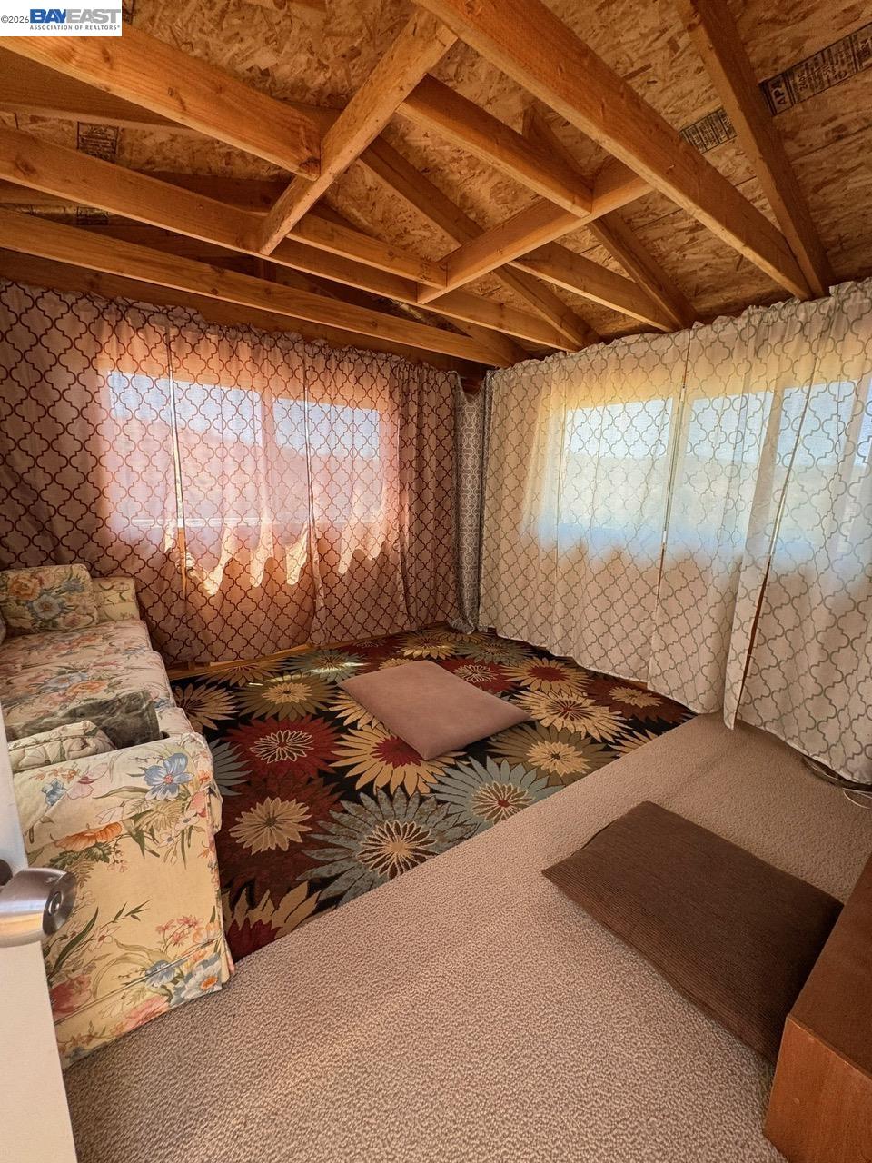 Furnished room with exposed wood rafters, patterned curtains, floral sofa, and colorful carpet.