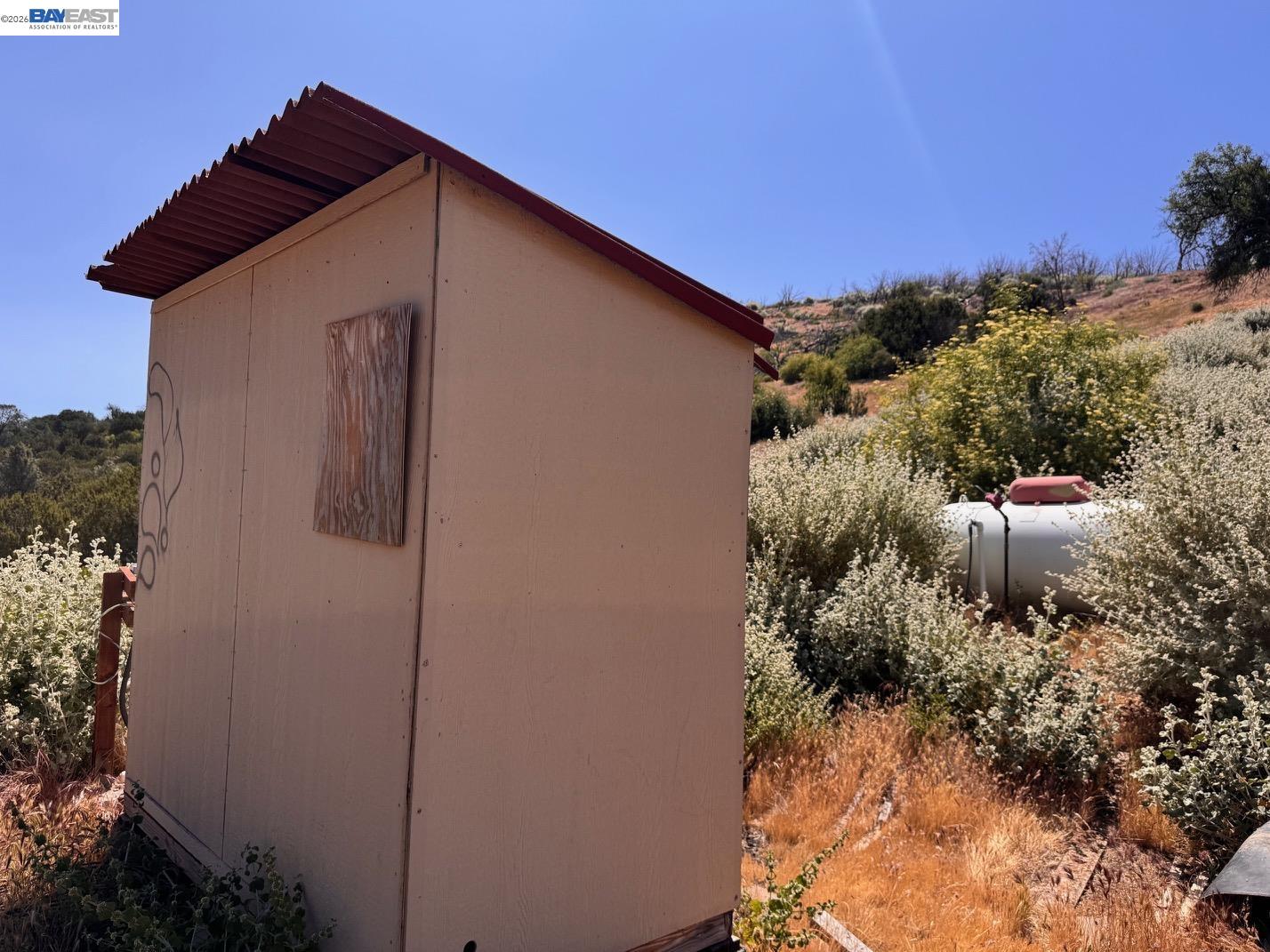 Small utility shed and storage tank amid dry brush on a rural hillside.