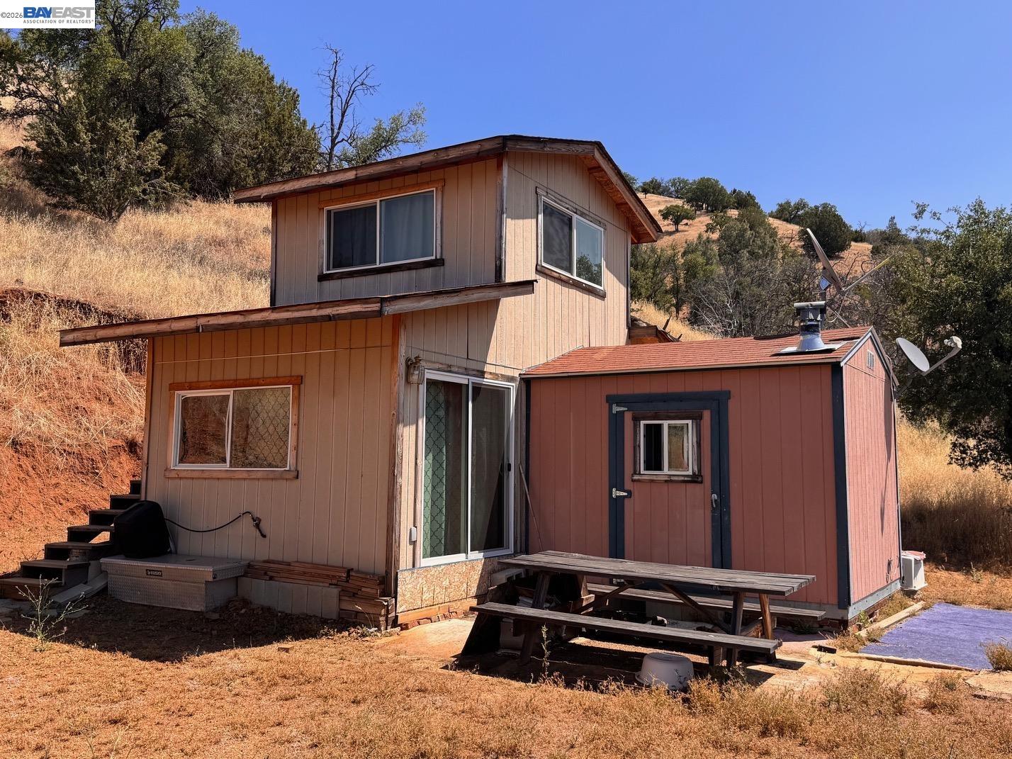 Small two-story home with attached red outbuilding on a dry, tree-dotted hillside.