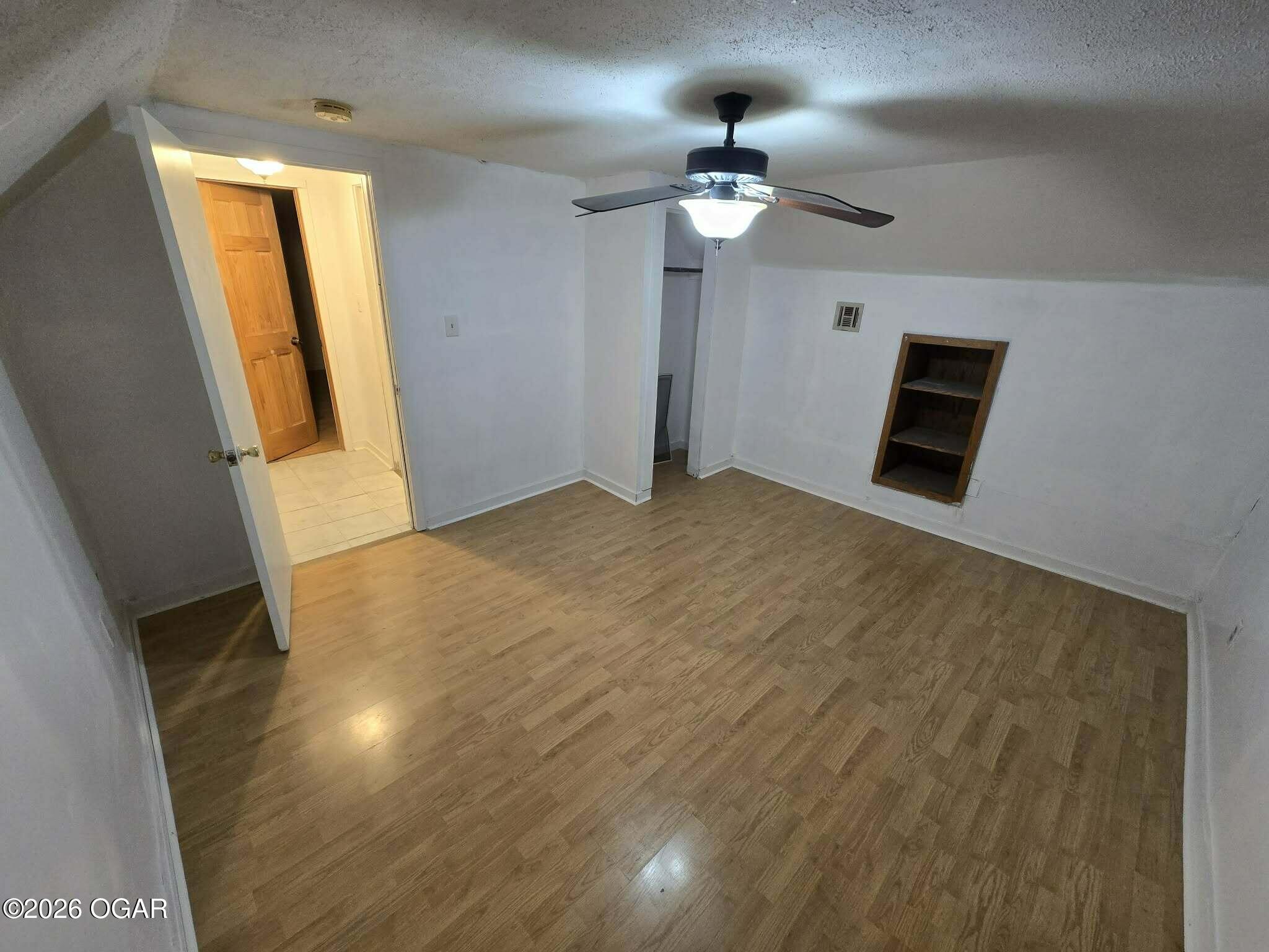 Empty room with wood-look flooring, ceiling fan, and built-in wall shelf beside an open doorway.
