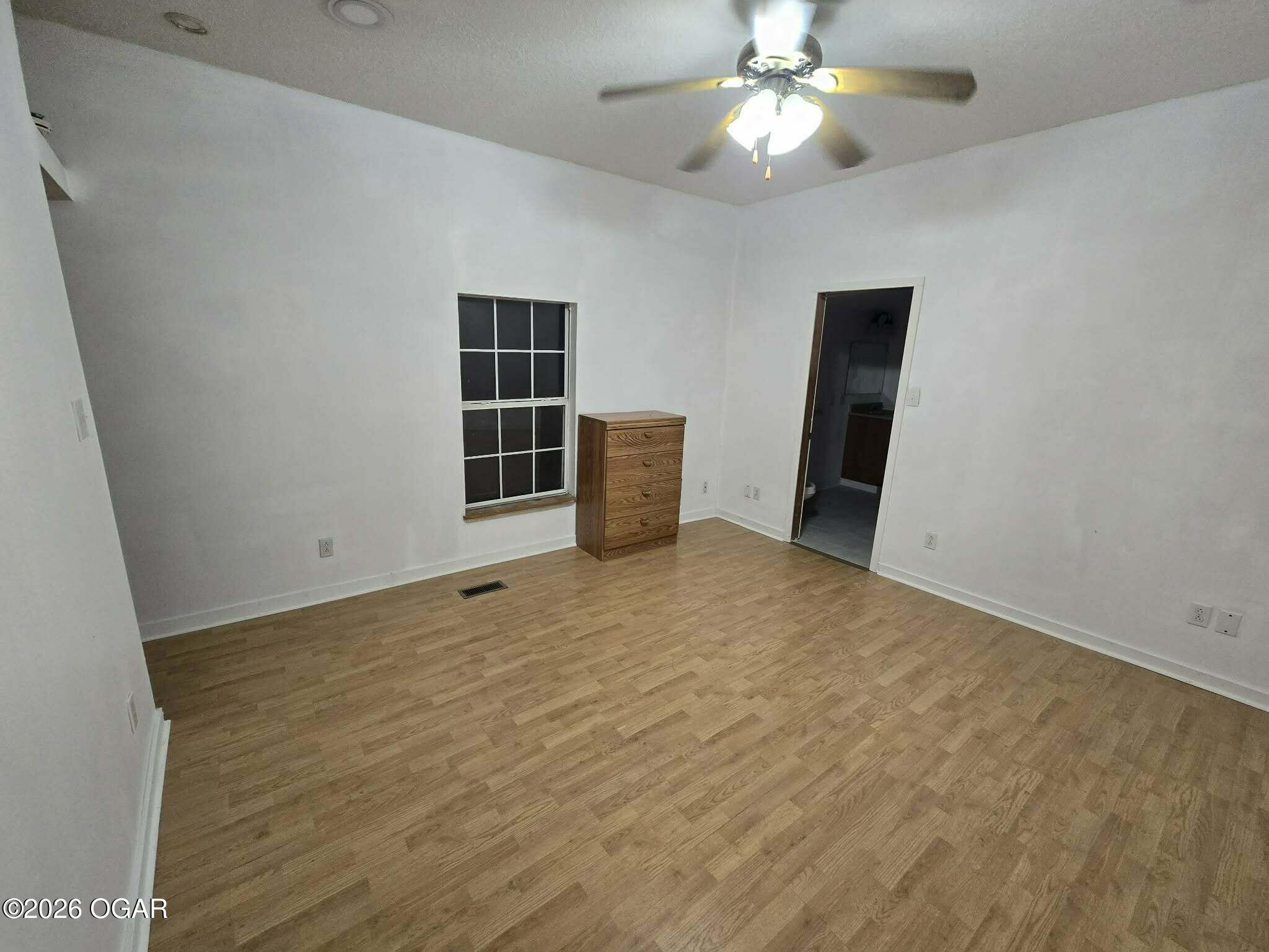 Bedroom with light wood floors, a ceiling fan, dark-paned window, and wooden chest of drawers.