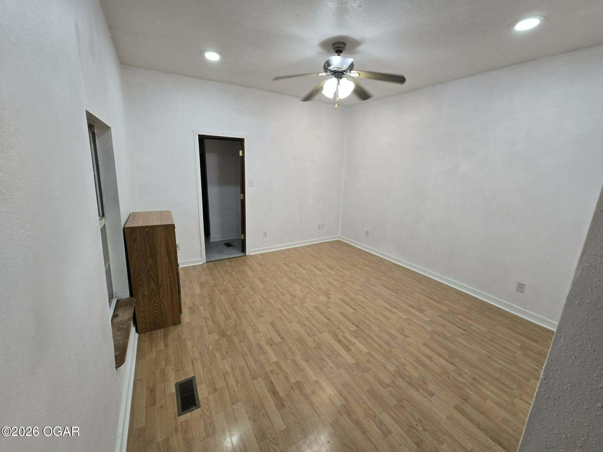 Mostly empty room with wood-look flooring, a ceiling fan, and a small cabinet beside the window.
