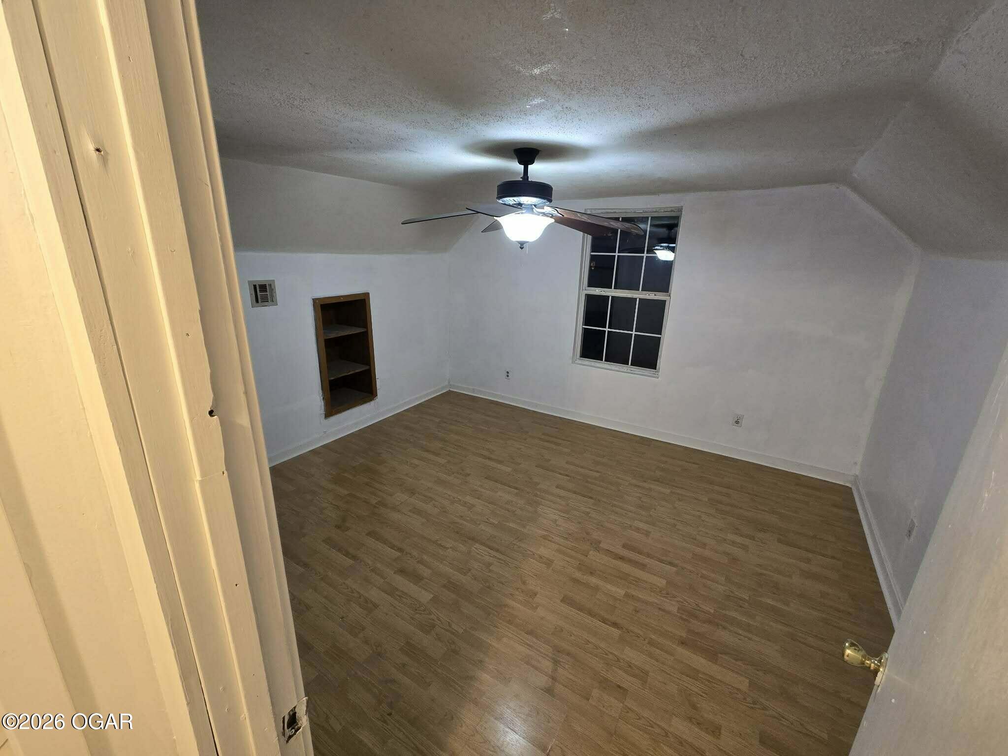 Empty room with wood-look flooring, a ceiling fan, a window, and a built-in shelf.