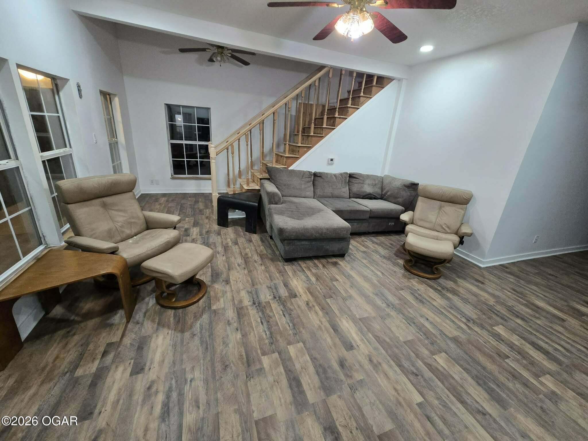Furnished living room with a gray sectional, tan recliners, wood-look flooring, and staircase.