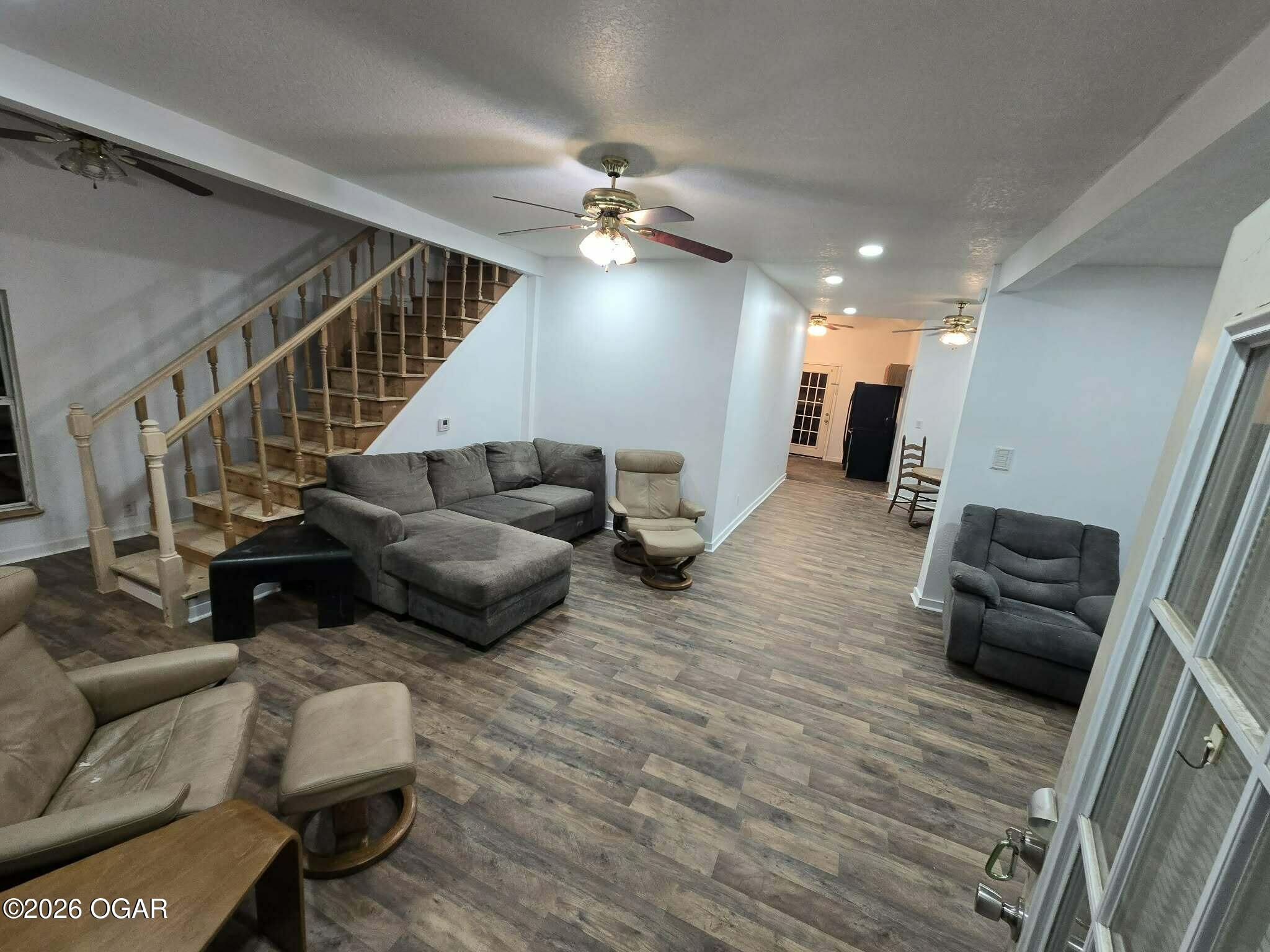 Living room with gray sectional, recliners, wood-look flooring, and a staircase.