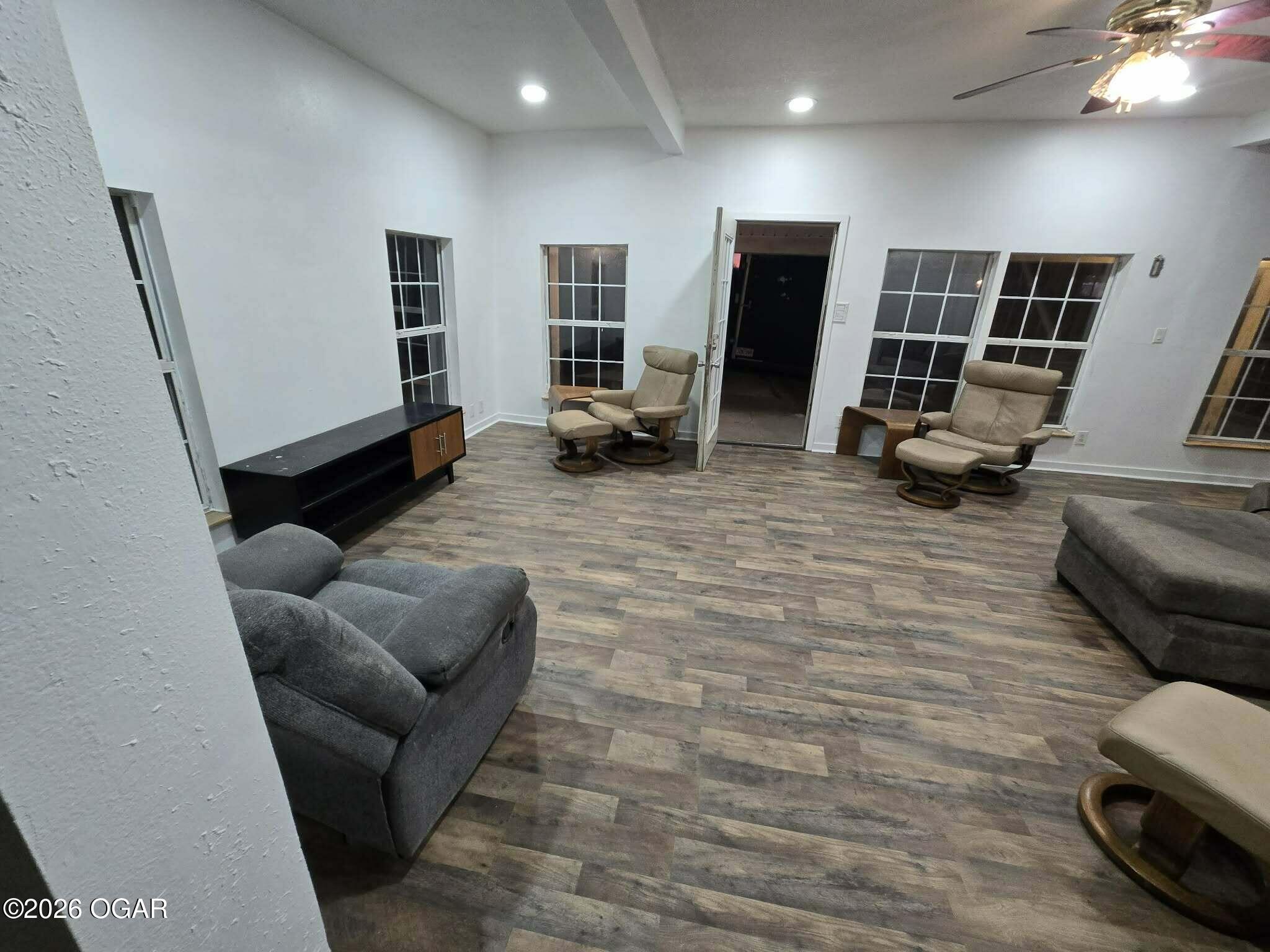 Furnished living room with wood-look flooring, multiple recliners, and windows beside an open doorway.