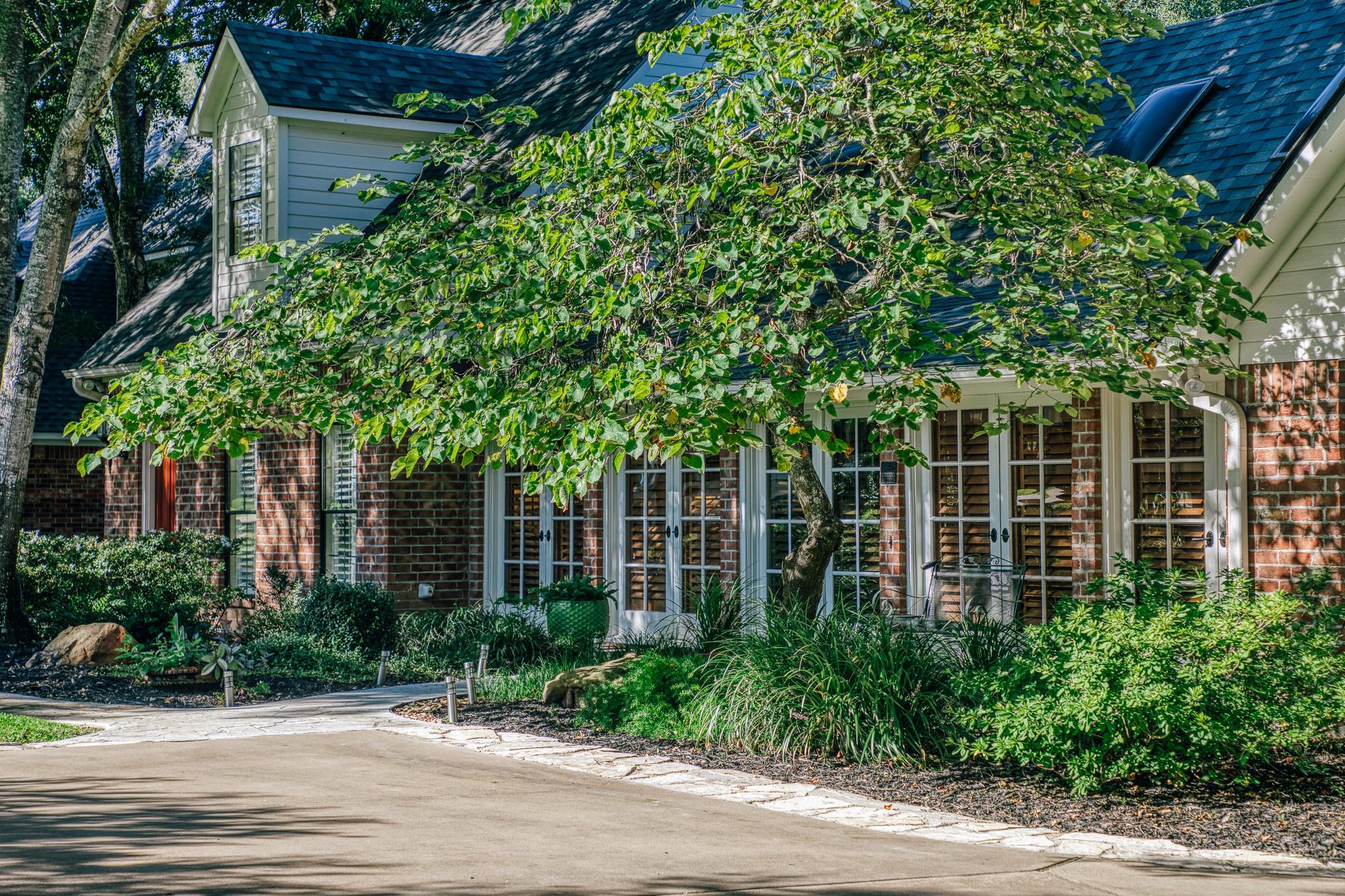 Two-story brick-and-siding house with French doors, shaded by a mature tree and surrounded by landscaping.