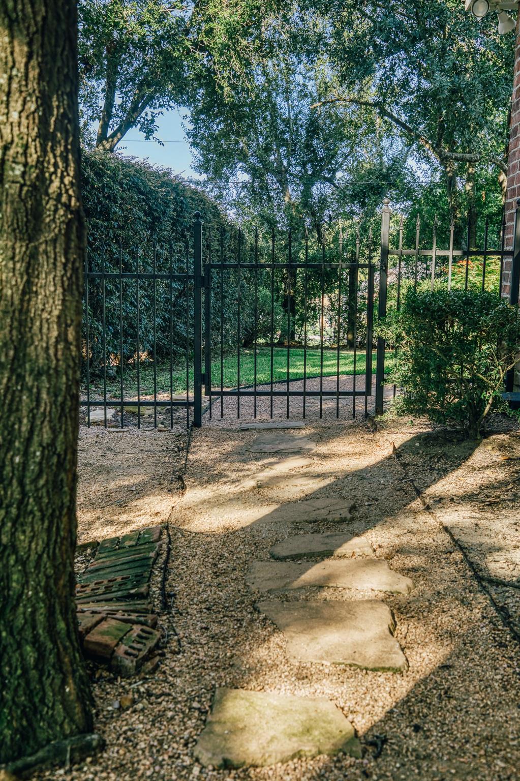 Garden path leading through a black metal gate, shaded by mature trees and bordered by hedges.
