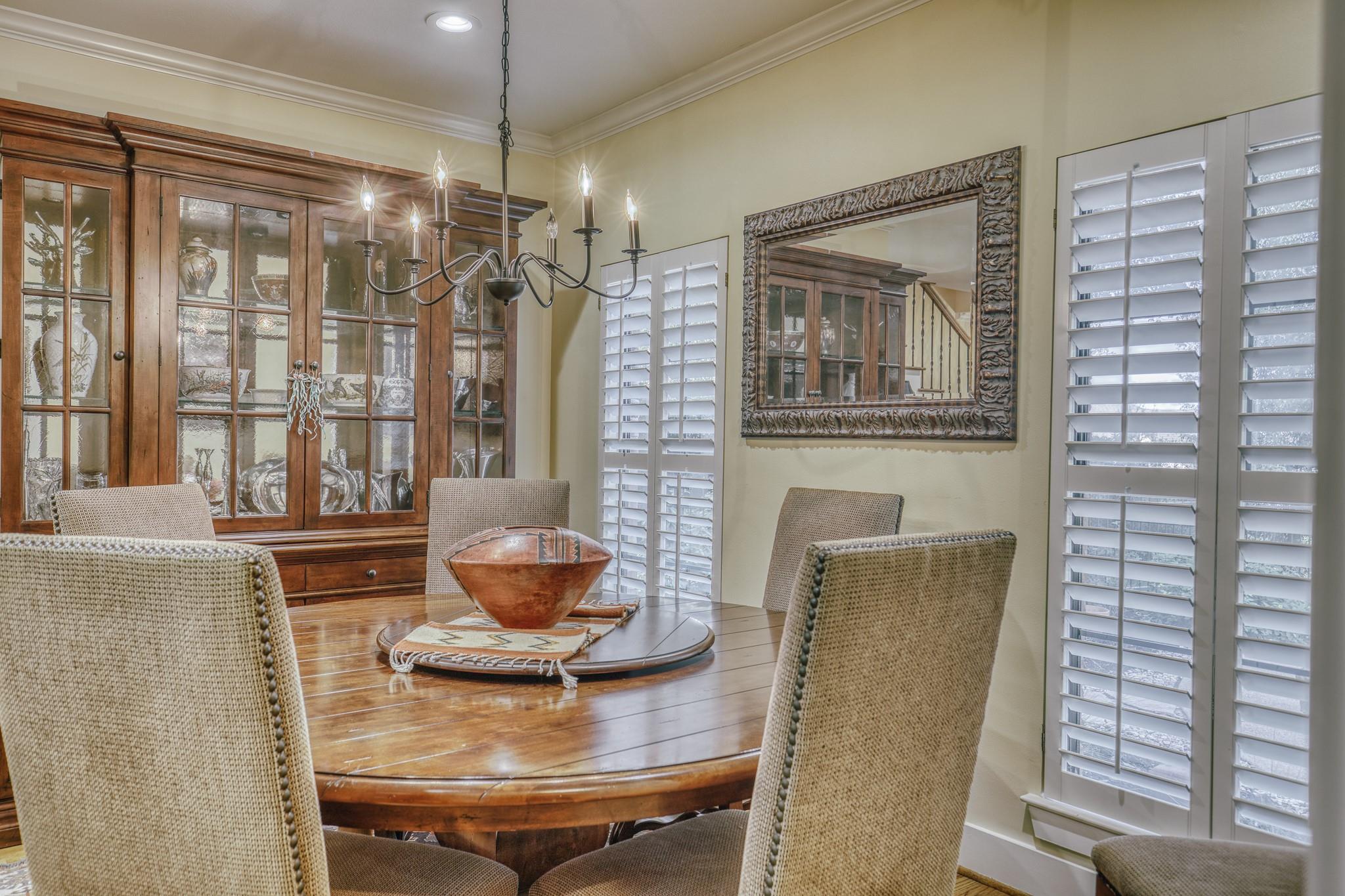 Dining room with an oval wood table, glass-front china cabinet, and white plantation shutters.